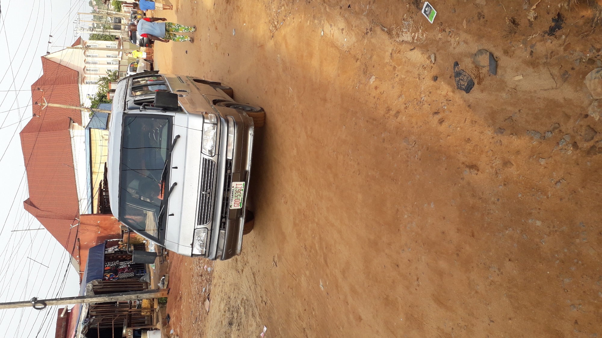 The image shows a silver Mitsubishi L300 van parked on a dirt road in Nkpor, Nigeria. The vehicle's license plate reads "GDD-536ZT." The road surface is dusty and uneven. Several people are visible in the background. A building with a red roof is visible. A market stall is seen in the background. Power lines are also visible above the scene.