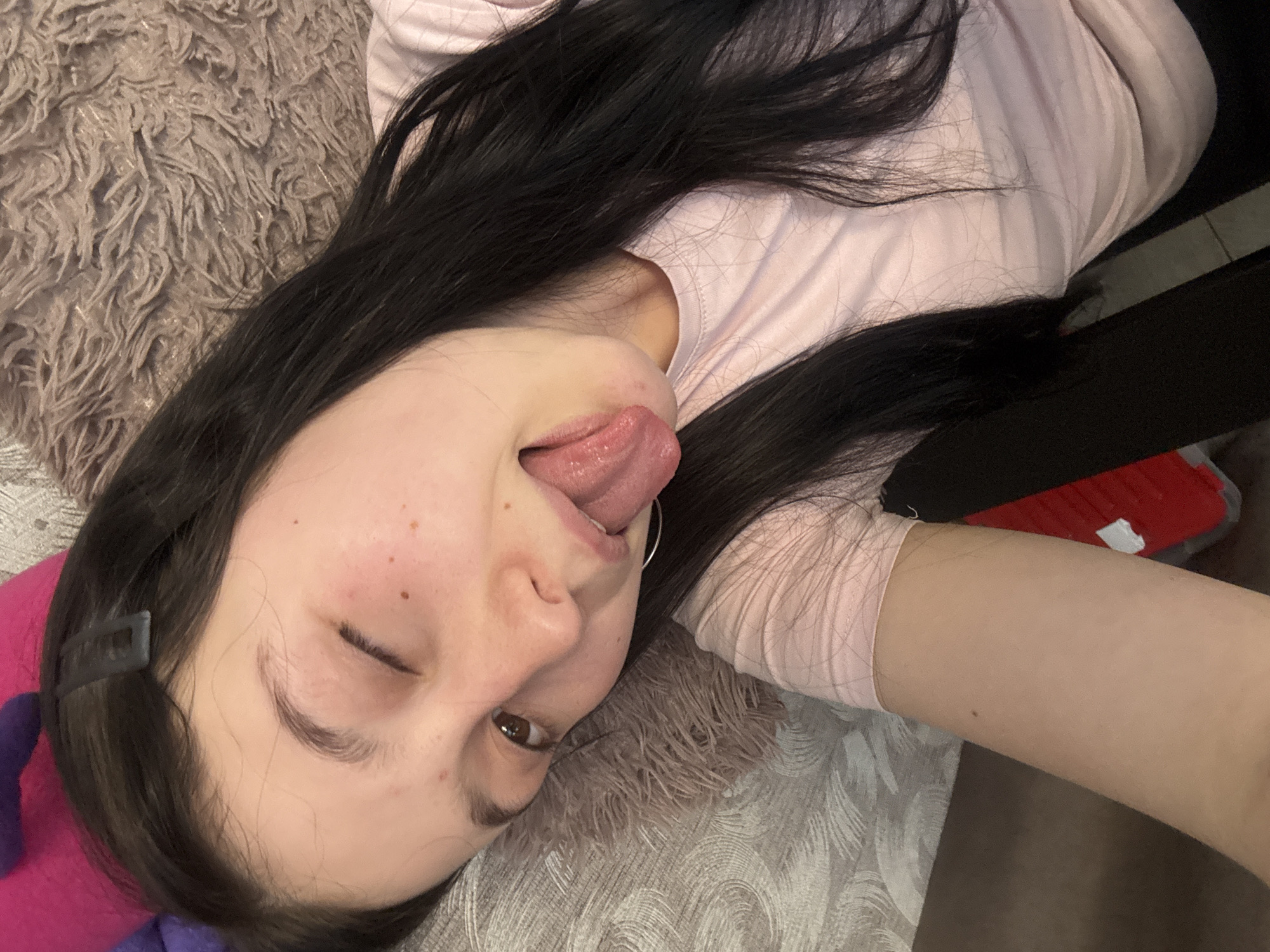 A woman with long, dark hair is lying down, sticking her tongue out. She is wearing a light pink shirt. She appears to be on a soft, textured surface. A red object is partially visible. There is no information in the image to determine a specific location in Фоки, Russia.