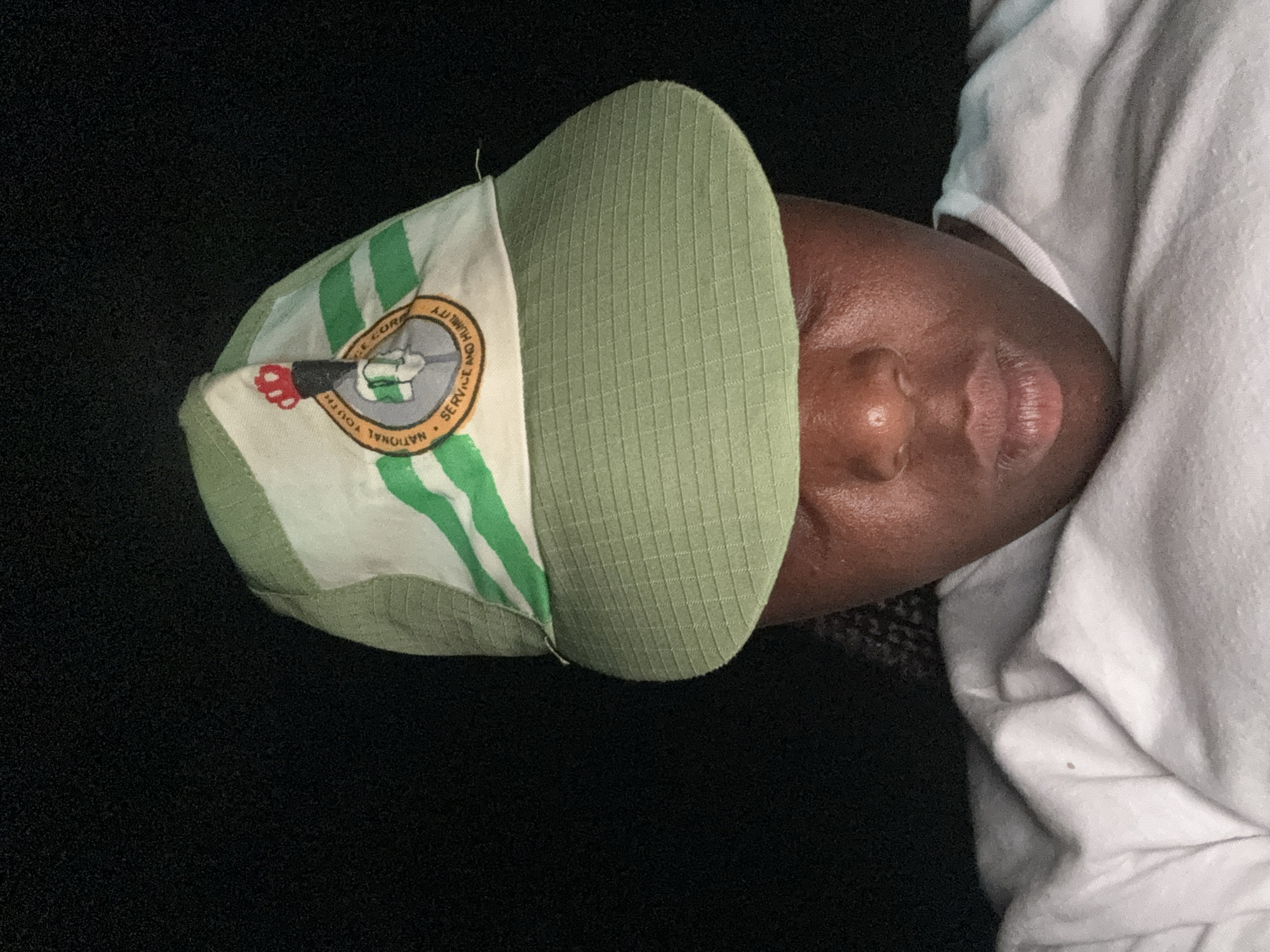 A close-up, portrait-style, rotated image shows a dark-skinned person wearing a green and white hat. The hat bears the inscription "National Youth Service Corps. Service and Humility." The person is lying on their back, wearing a white shirt. The background is black. No specific location or city details are discernible from the image.
