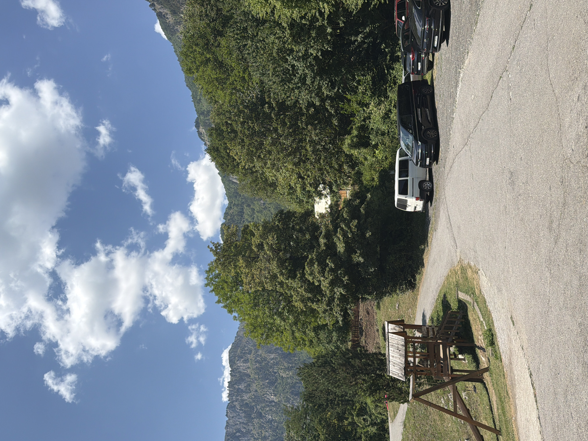 A dirt and asphalt road curves through a green, mountainous area. A wooden playground structure stands on the left side of the road. Several vehicles, including white and black vans and SUVs, are parked on the right side of the road. The background is dominated by lush green trees and rocky mountain slopes. The sky above is blue with scattered white clouds.