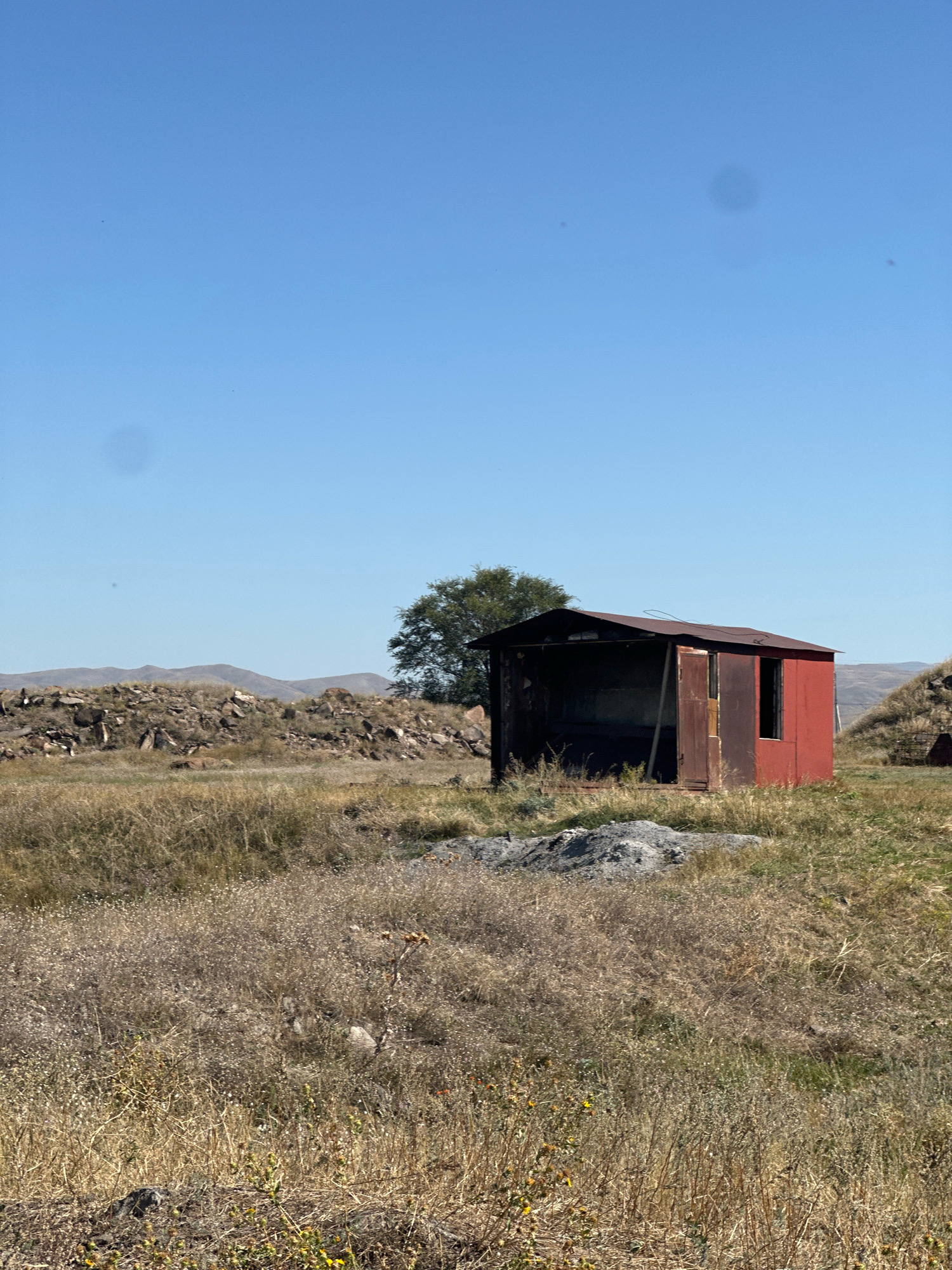 The image depicts a small, weathered red shed with a rusty metal roof, set in a dry, grassy field under a clear blue sky. The shed has an open front and a closed door on its side, with a window also visible on the side. A pile of grey gravel sits in front of the shed. In the background, a solitary tree stands to the left of the shed, and rolling hills are visible in the distance. The overall environment appears arid and rural, with sparse vegetation and rocky terrain. It is difficult to ascertain the precise location within Gyumri, Armenia, as no identifying landmarks are present. The image captures a still, daytime scene, with no people or animals visible.