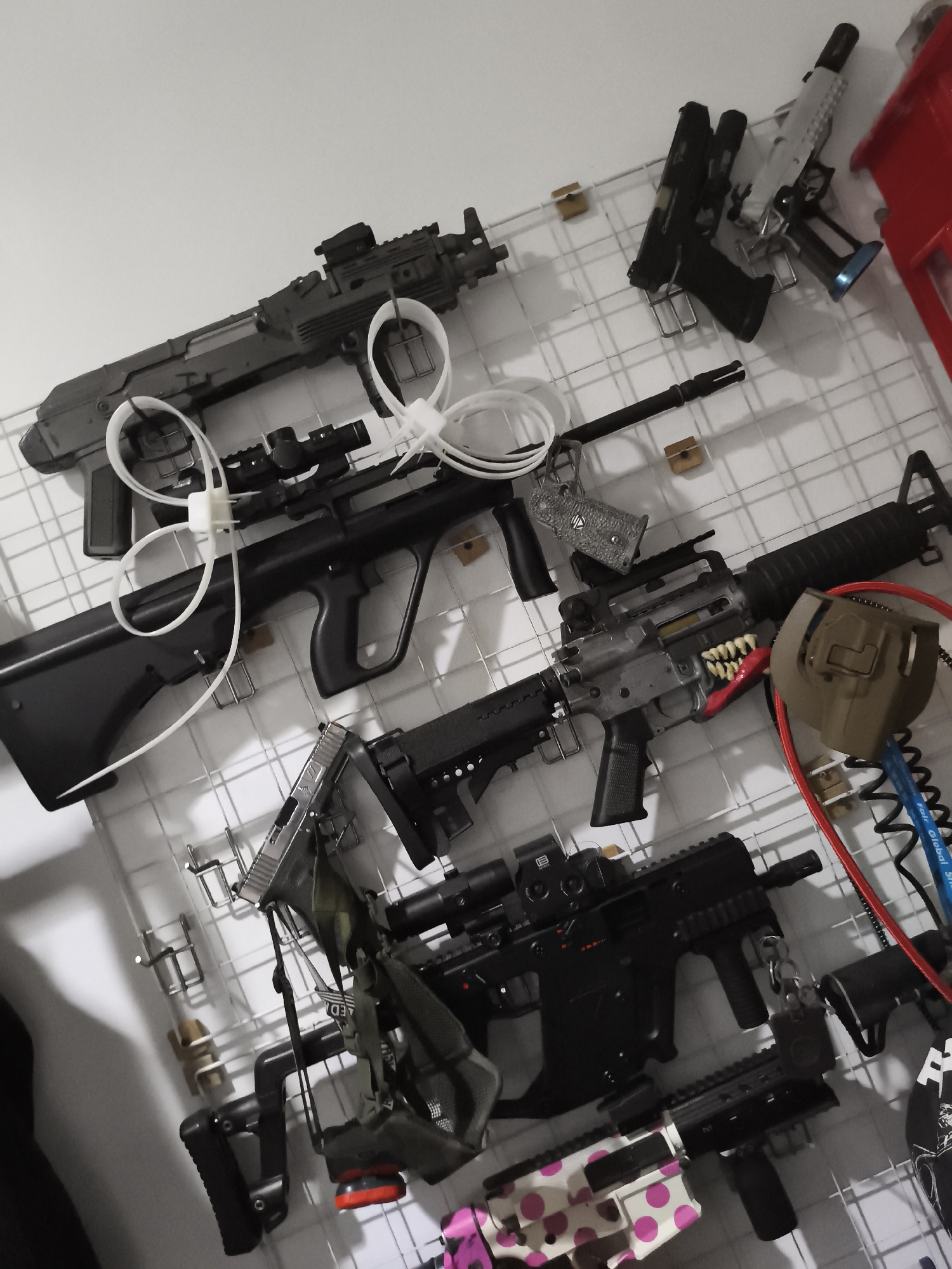 In West Jakarta, Indonesia, an indoor scene displays a collection of replica firearms, likely airsoft guns, mounted on a white grid wall. The firearms are arranged in rows, with several long rifles and pistols visible. Some of the guns are secured with white zip ties or placed on metal hooks. One rifle features a distinctive "fang" design around its barrel, and another has a camouflage pattern with pink polka dots. Several accessories, including scopes, grips, and holsters, are also attached to or displayed near the firearms. The lighting in the room is dim, casting shadows that add to the overall atmosphere.