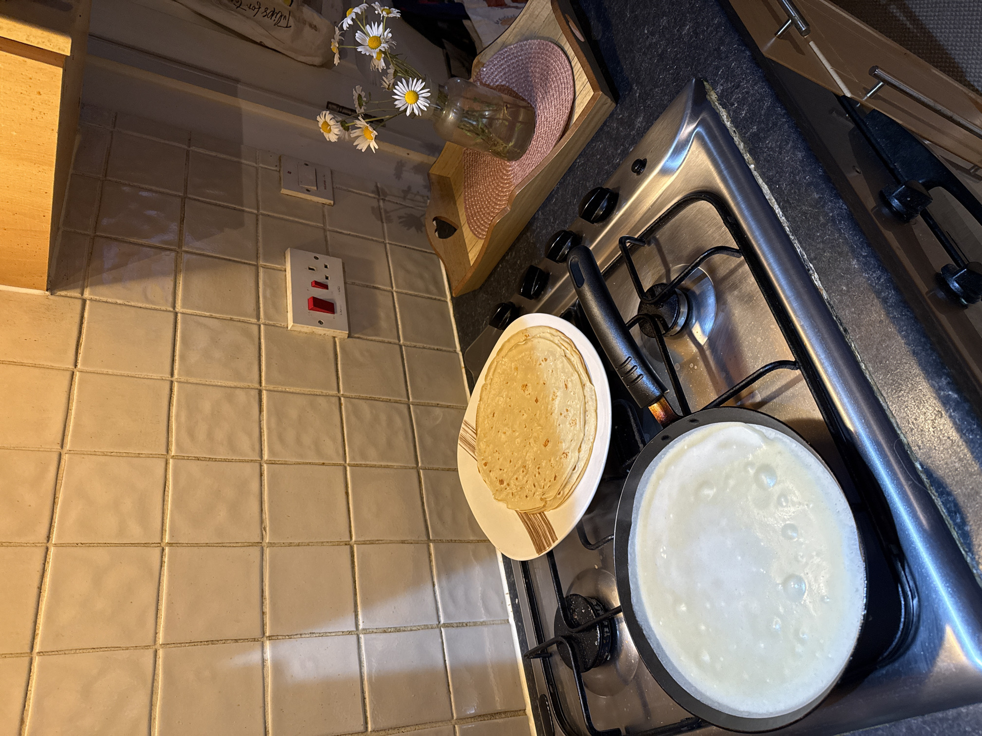 The image depicts a domestic kitchen scene, likely in Great Yarmouth, United Kingdom, featuring a gas stovetop where pancakes are being made. A stack of cooked pancakes rests on a plate beside a frying pan filled with uncooked batter. A vase of daisies sits on a wooden tray to the left of the stovetop. The kitchen backsplash consists of square ceramic tiles. A UK standard electrical outlet is visible on the tiled wall.