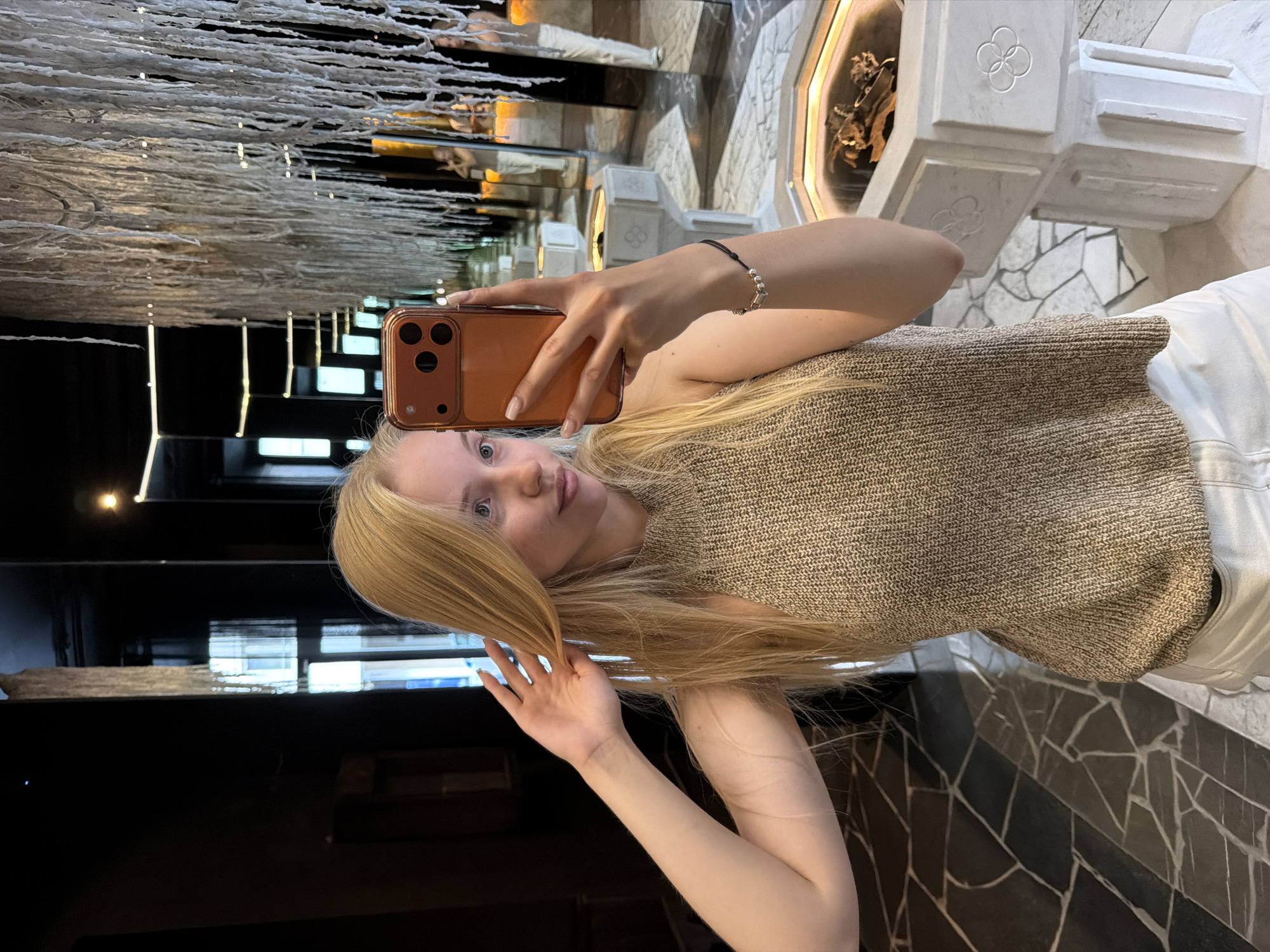 A young blonde woman takes a mirror selfie in an indoor setting. She holds an orange-brown smartphone with three visible camera lenses in her right hand, positioned to capture her reflection. Her left hand is raised, lightly touching her blonde hair near her left ear. She is wearing a light brown/tan sleeveless knitted top with a mock neck and white trousers. A black bracelet with silver bead details is on her right wrist. The background, largely composed of reflective surfaces, creates an illusion of a deep, mirrored hall. Above, a prominent art installation of numerous white, string-like textured elements hangs from the ceiling. The floor features an irregular pattern of dark and light gray stone tiles. To the woman's right, a white, multi-faceted pedestal or display stand with carved four-leaf clover-like patterns is visible, containing dark, organic material under a clear surface. Further into the reflections, similar pedestals are seen, along with reflections of other individuals in the distance and a tall window structure revealing glimpses of a city outside.