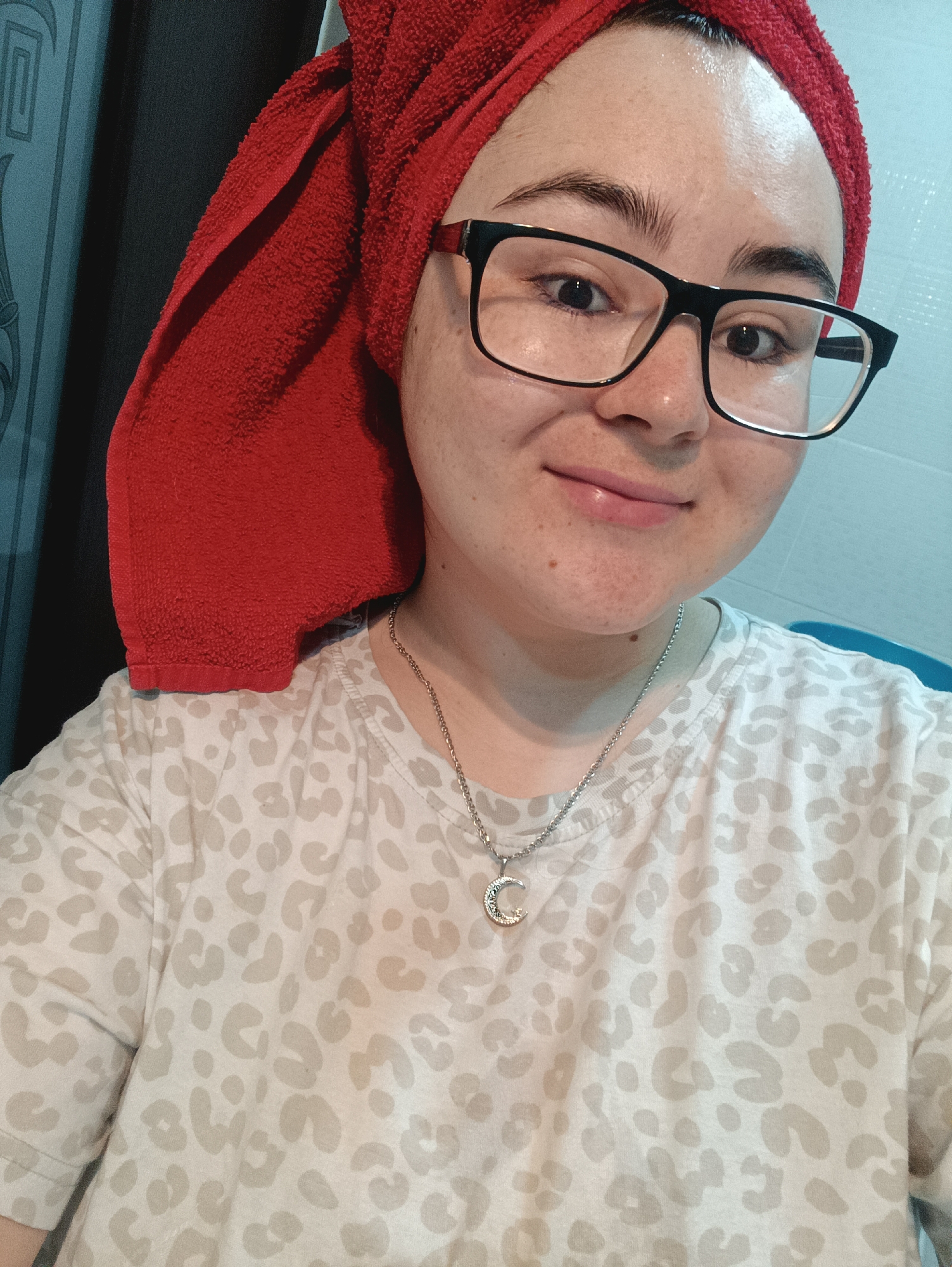 A young person is shown in a selfie. The individual has a red towel wrapped around their head and is wearing black-framed glasses. They have fair skin and a small, crescent moon necklace. The person is wearing a white t-shirt with light gray animal print spots. The background appears to be a bathroom setting with blue-grey paneling. The location in the city of Noviy, Russia cannot be verified from the image.
