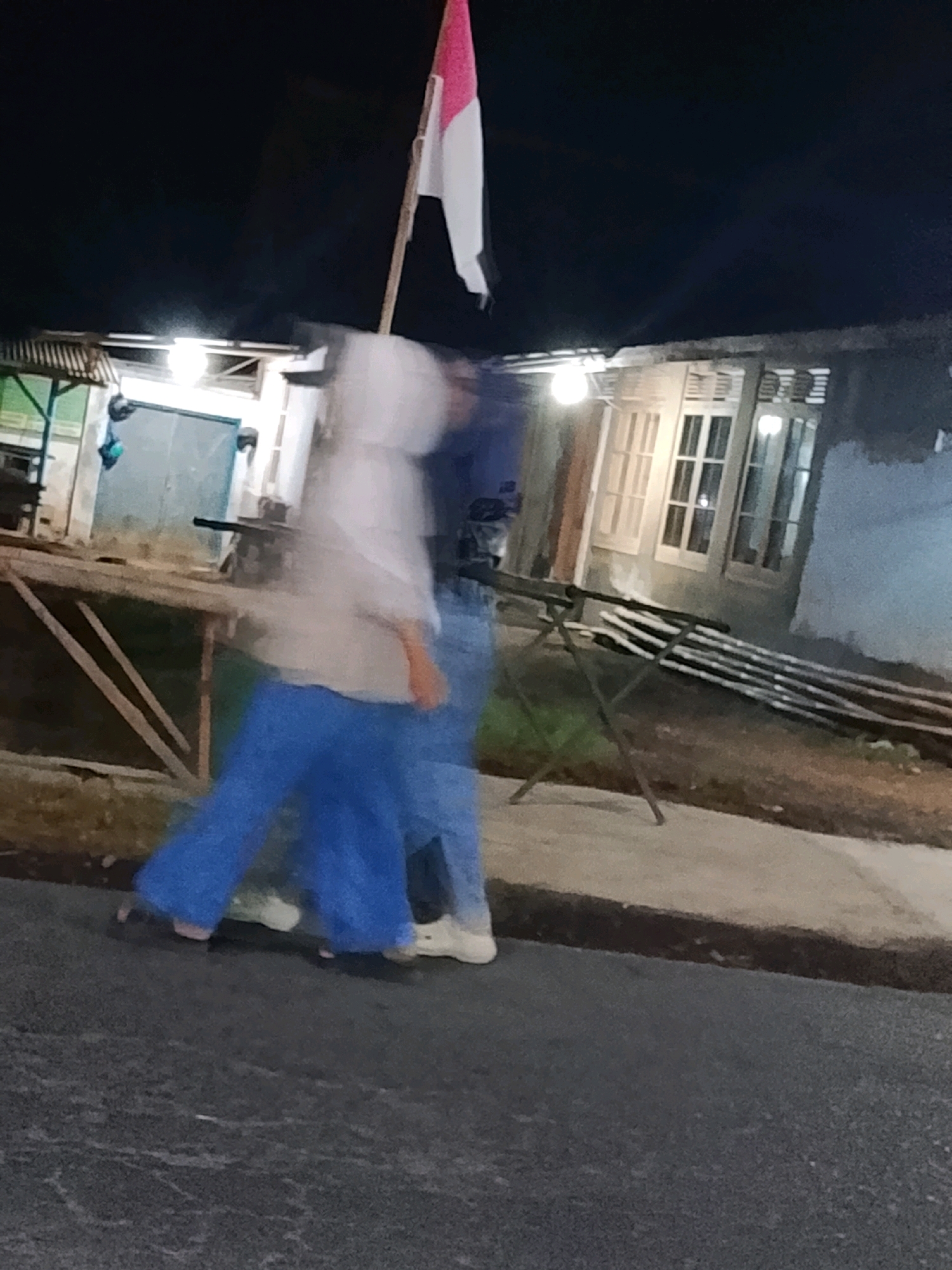 In Sukorejo, Indonesia, at night, a person wearing a white hooded jacket and blue pants is walking on a paved surface. They are holding a flag on a pole. Another person, blurred, is walking beside them. The background shows a building with windows and a roof, and some outdoor elements such as a table and poles supporting what appears to be stored materials. The scene is illuminated by artificial lights.