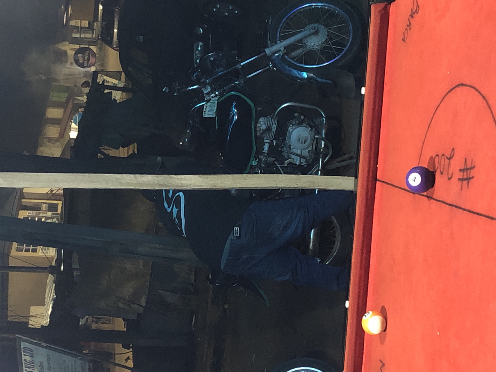 A man in blue jeans and a dark shirt with blue markings is bending over a black motorcycle in a dimly lit urban environment. The motorcycle has a license plate that reads "JJN 94Z". In the foreground, a red pool table is visible with two pool balls: one is a white and orange striped ball, and the other is a purple ball with the number "1" on it. The scene appears to be outdoors, possibly at night, with some ambient light sources reflecting off the motorcycle and the pool table.