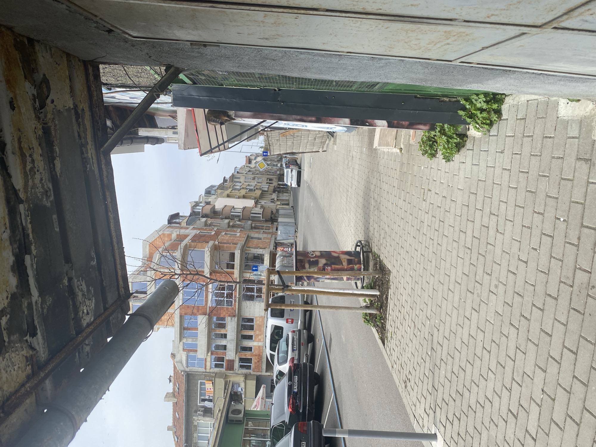 The image captures a street scene in Burgas, Bulgaria. A newly constructed building with brick facade and multiple windows is visible. Vehicles, including a white van and several dark-colored cars, are parked along the street. The street is paved with grey bricks and lined with trees in small planters. The perspective is from under an awning, obscuring the top left of the frame. Overhead wires and awnings extend over the street, and in the distance, more buildings are visible.