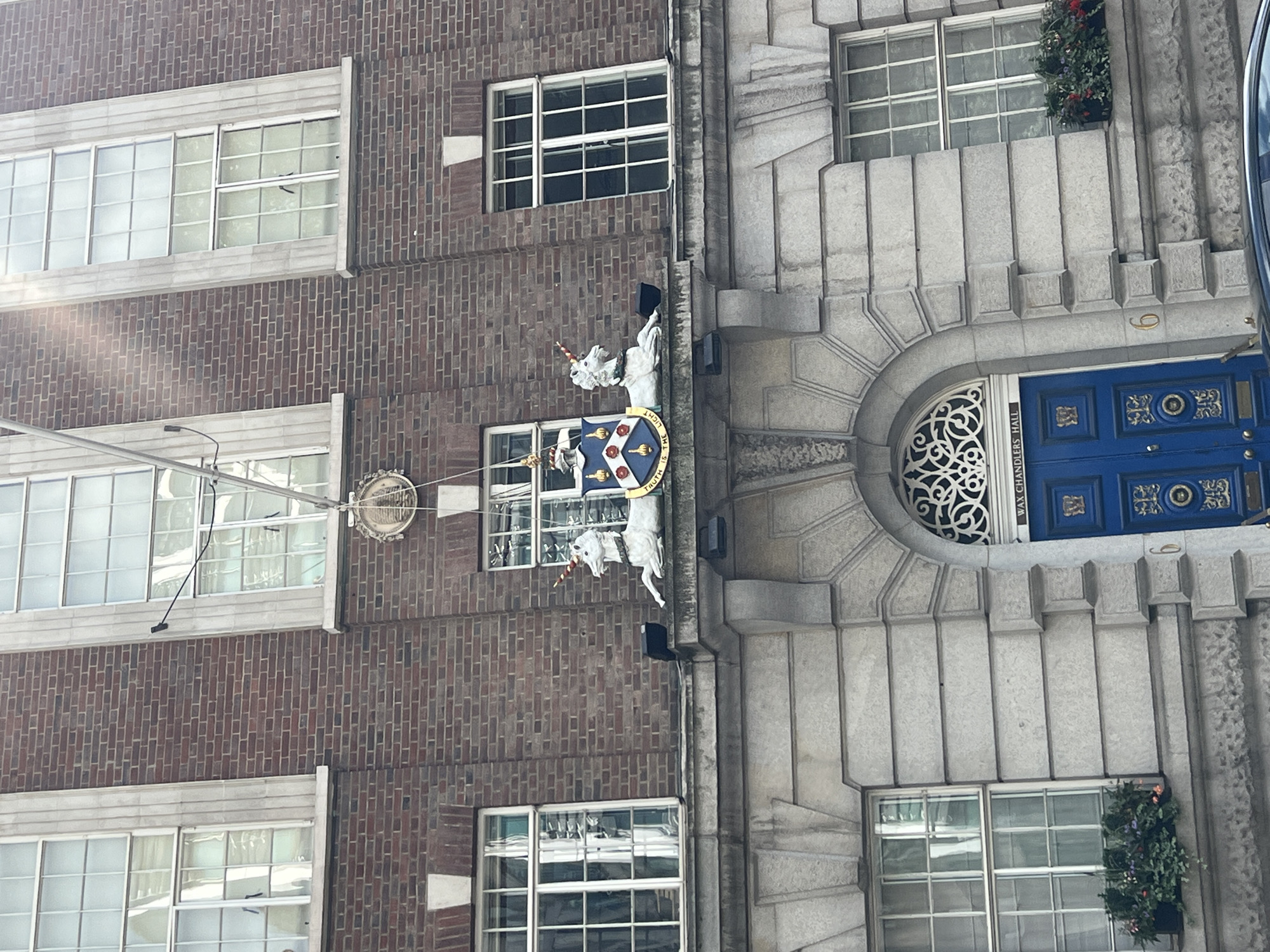 The image shows the exterior of a building in the City of London, United Kingdom. The building is constructed of red brick and features large, multi-paned windows. Above the entrance, there is a stone facade with decorative carvings, including two white unicorns and a coat of arms. The entrance itself is a large blue door with gold ornamentation, situated beneath an arched window with intricate metalwork. Above the door, a sign reads "WAX CHANDLERS HALL". To the left and right of the entrance, there are windows with flower boxes filled with green foliage and red flowers. The overall impression is one of historic architecture and traditional design.