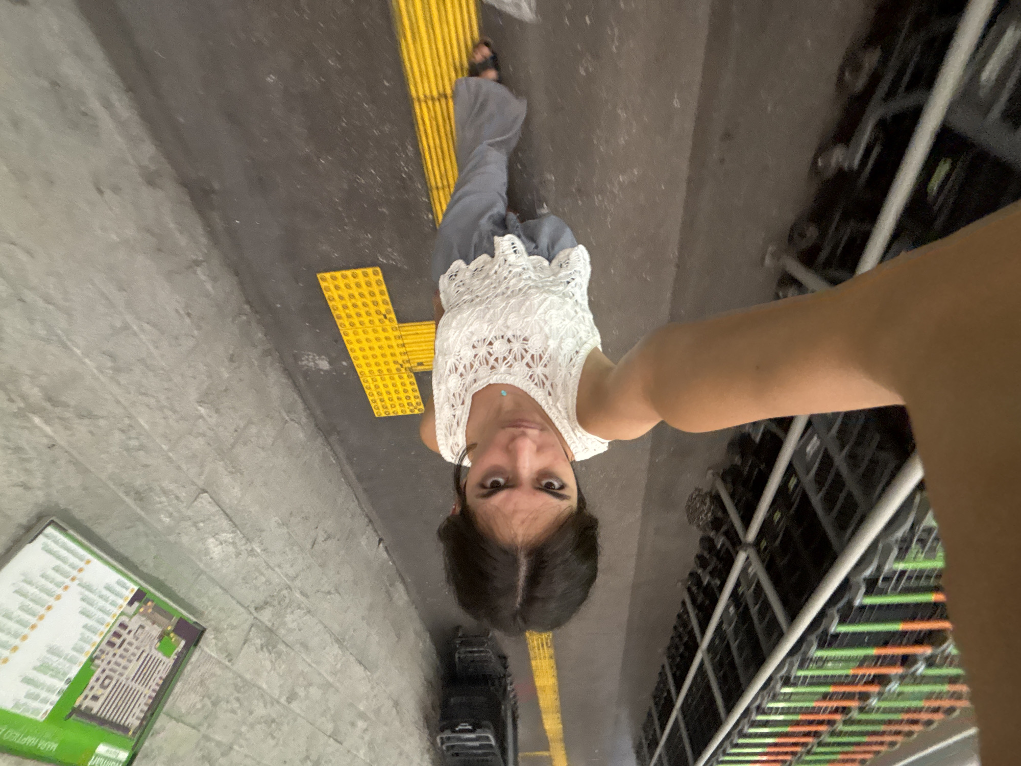 A tilted, upward-facing selfie captures a person standing on a walkway marked with yellow tactile paving. The person wears a white crochet top, grey pants, and sandals. To the right are rows of shopping carts stacked together. To the left is a wall with a map of the location, identified as a Walmart store. The city is Puebla, Mexico.