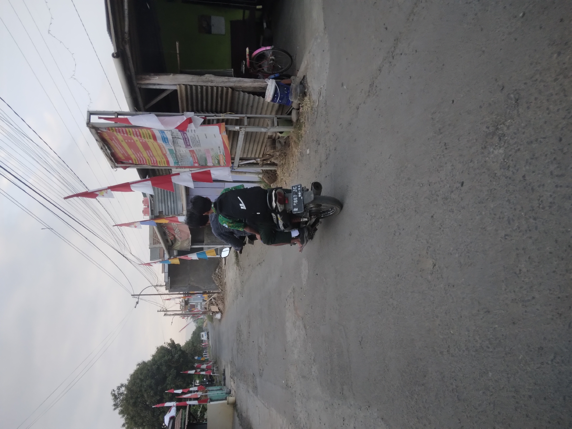 The image depicts a street scene in Desa Pamijen Kecamatan Sokaraja, Indonesia, with a focus on a section of asphalt road lined with buildings and Indonesian flags. Two individuals are riding a motorcycle, with the person in the rear wearing a backpack. The motorcycle's license plate is partially visible. Various flags, predominantly red and white, are displayed along the roadside, suggesting a festive or patriotic occasion. Buildings with corrugated metal roofs and walls are present, some appearing to be shops or residences. A bicycle is parked to the right of the scene. Electrical wires are strung overhead, a common sight in urban and semi-urban environments. The sky is overcast, indicating diffused lighting conditions. The road surface is weathered and shows signs of wear.