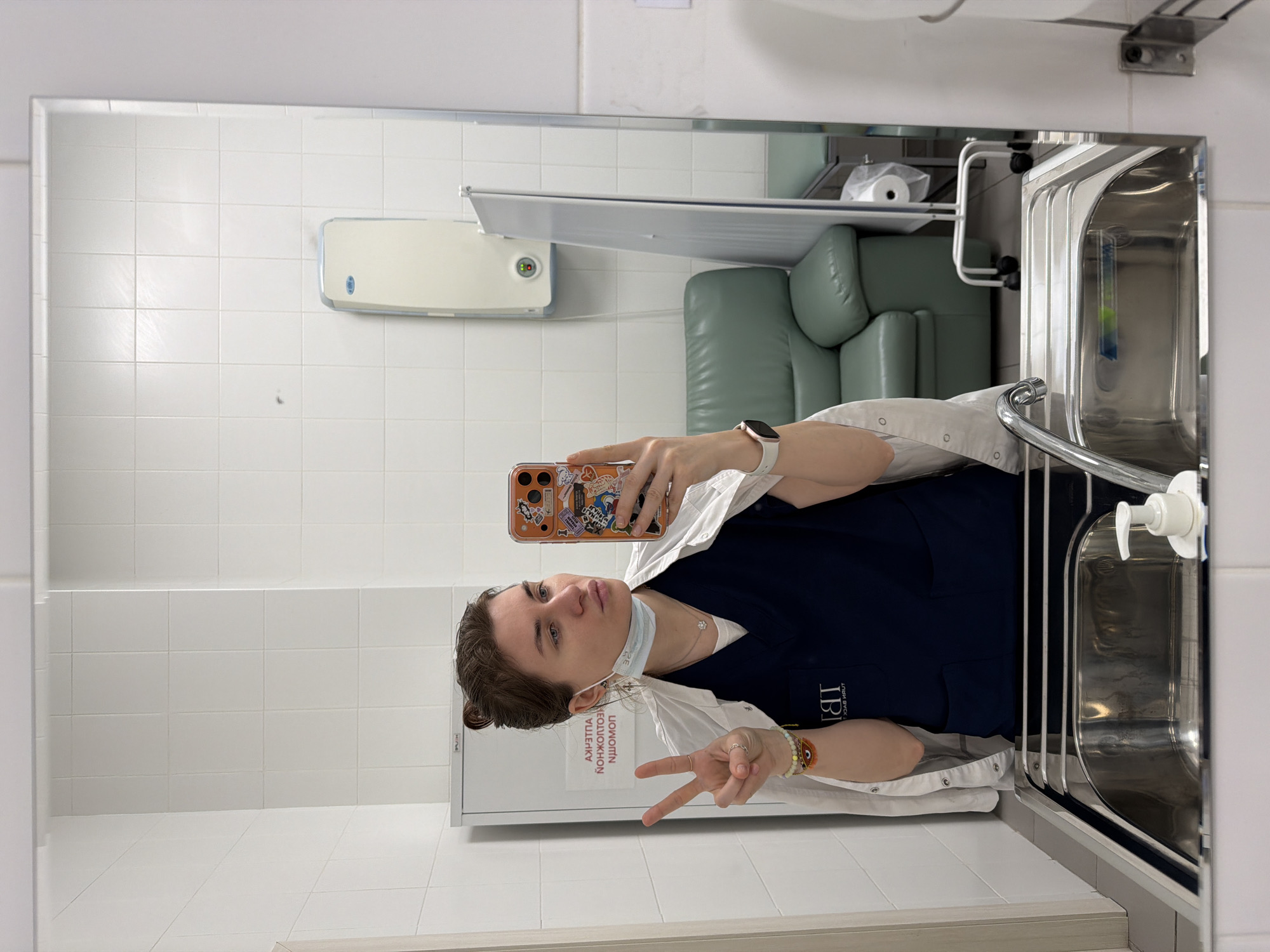 A person is taking a mirror selfie in what appears to be a clean, tiled room, possibly within a medical facility or public space. The individual, a woman, is holding a smartphone with a colorful, stickered case, making a peace sign with her left hand. She is wearing dark blue medical scrubs with a white overshirt, a medical mask pulled down to her chin, a smartwatch on her left wrist, and beaded bracelets on her right wrist, one featuring an evil eye charm. Her hair is pulled back.
The reflection in the mirror reveals a bright, sterile environment with white tiled walls and floor. In the background, there is a baby changing station mounted on the wall, and to its right, a light green armchair or sofa. Further right, a metal sink with a faucet and a white pump soap dispenser is visible. Above the sink, a shelf holds a roll of paper. Behind the person, out of the mirror's reflection, is a white cabinet.
Visible text on the cabinet, which appears reversed in the selfie, reads "СМЕНА ПЕЛЕНОК" in Cyrillic, meaning "DIAPER CHANGE". On the person's scrubs, a reversed logo "IB" is partially visible, likely part of an institutional abbreviation. The person's expression is neutral to slightly smiling, conveying a casual mood. The location context provided is Khimki, Russia.