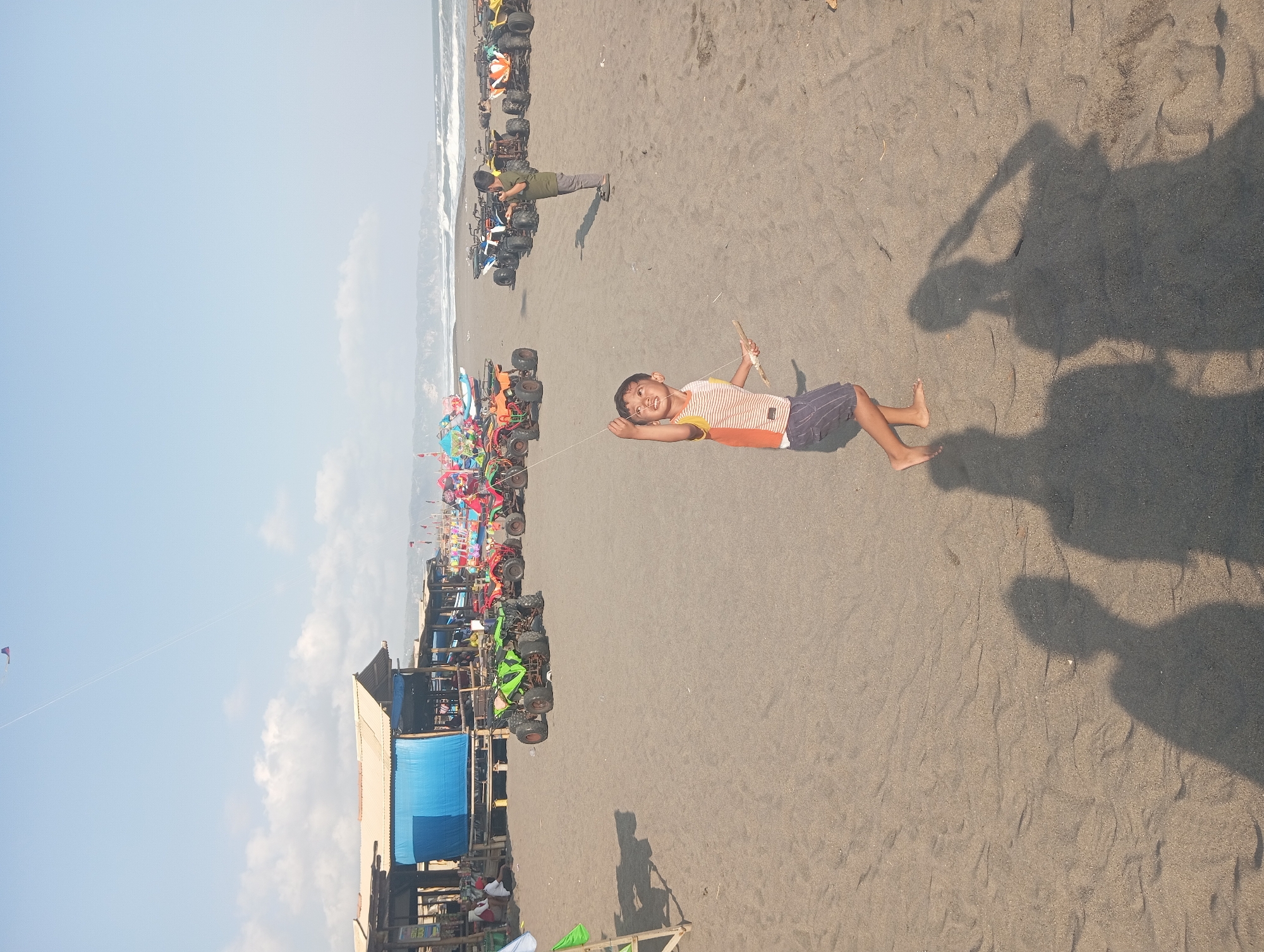 In Parangtritis, Indonesia, a young boy is depicted on a sandy beach, actively flying a kite. He is positioned in the foreground, facing upwards and holding the kite string with both hands. The boy is wearing a striped shirt and dark shorts. Behind him, to the right, another boy stands, also looking upwards. A line of colorful ATVs is parked on the sand further back, leading towards a row of wooden stalls with blue awnings. The ocean is visible in the background with waves rolling onto the shore, and the sky is a clear blue with scattered clouds. The overall scene captures a typical day at the beach with recreational activities.