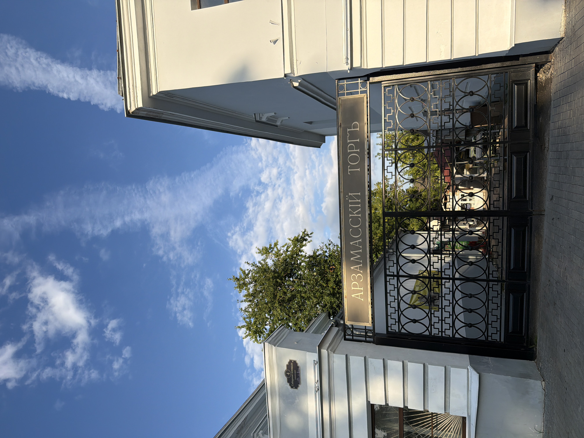 The image captures an ornate black metal gate in front of a building, under a blue sky with scattered white clouds. The gate has a decorative top with a sign that reads "АРЗАМАССКИЙ ТОРГЪ" in Cyrillic letters. The building to the left of the gate is light grey with white decorative elements. Above the gate, a darker sign with the number "4" and Cyrillic text is visible on the building's facade. A large green tree stands behind the gate. The ground in front of the gate is paved with rectangular stones. The overall impression is a historic architectural facade. The sign clearly indicates that this location is in the city of Arzamas, Russia, and is related to commerce or trade ("ТОРГЪ").