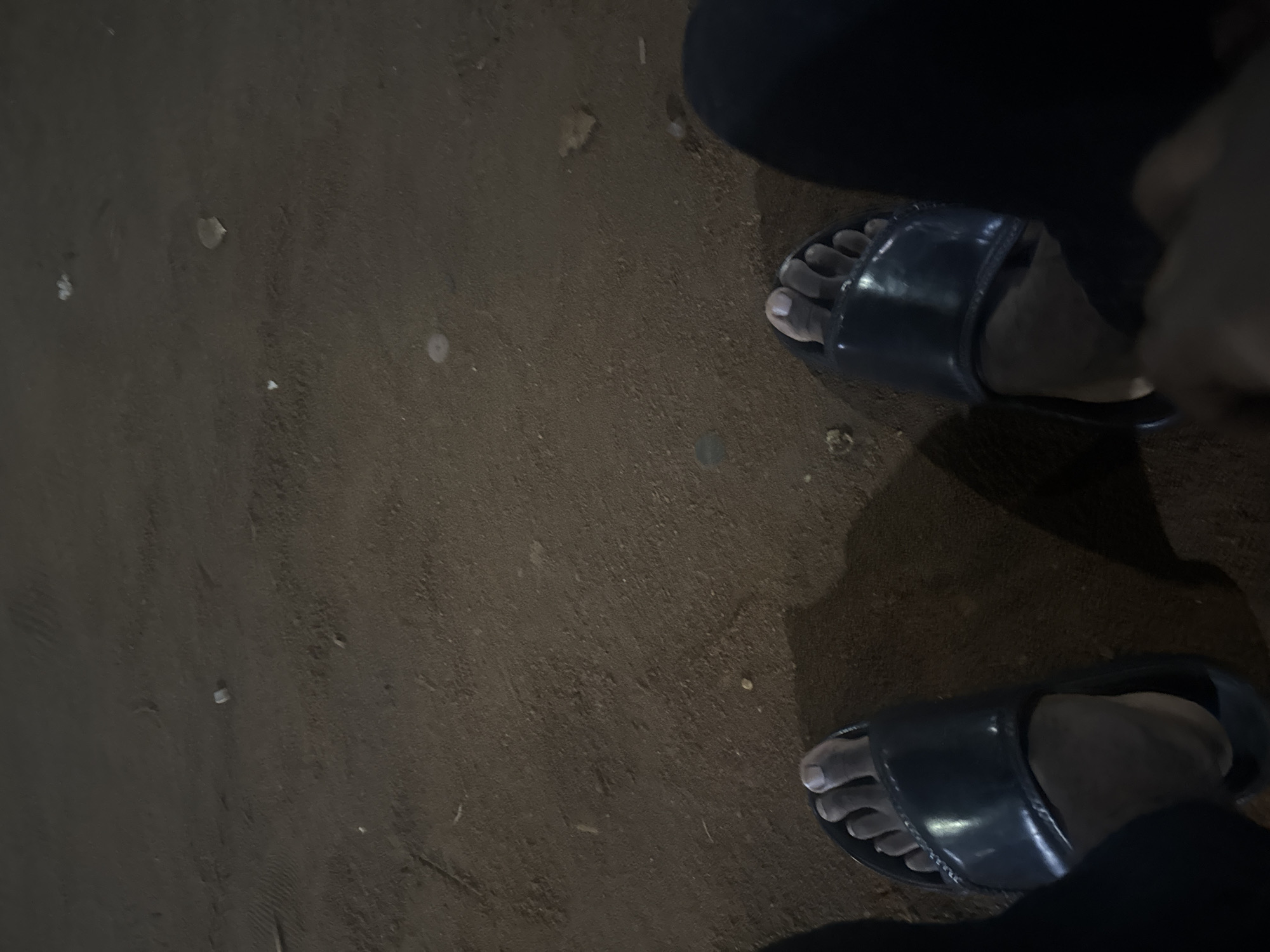 The image shows two feet wearing dark-colored slides on a dirt-covered surface. The location is possibly in Ihiagwa, Nigeria. The person is wearing dark clothing.