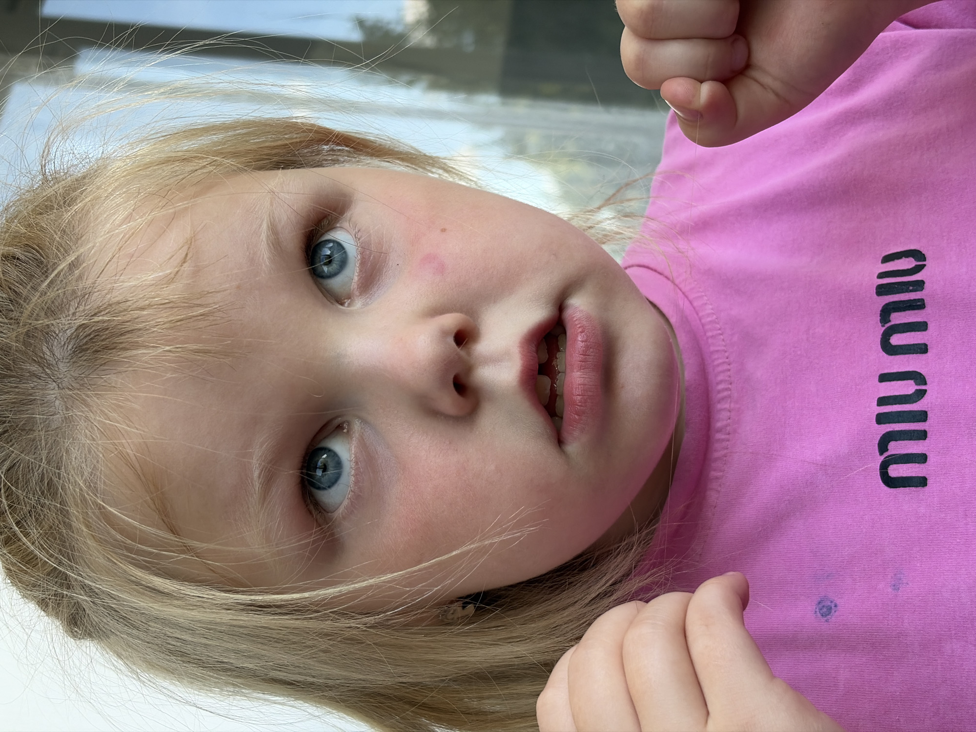 A close-up, waist-up, medium shot captures a young girl with blonde hair and blue eyes. She is wearing a bright pink t-shirt with "MIU MIU" in black lettering. The girl's mouth is slightly open, revealing her teeth, and her eyes are looking upwards. Her hands are raised, and she is holding a strand of hair between her fingers. There is a faint purple mark on her t-shirt. The background is blurred, suggesting an outdoor setting with a hint of greenery and a window. The image is a portrait with a shallow depth of field, focusing on the girl's face and upper body. The photograph was taken on a sunny day, as indicated by the lighting.