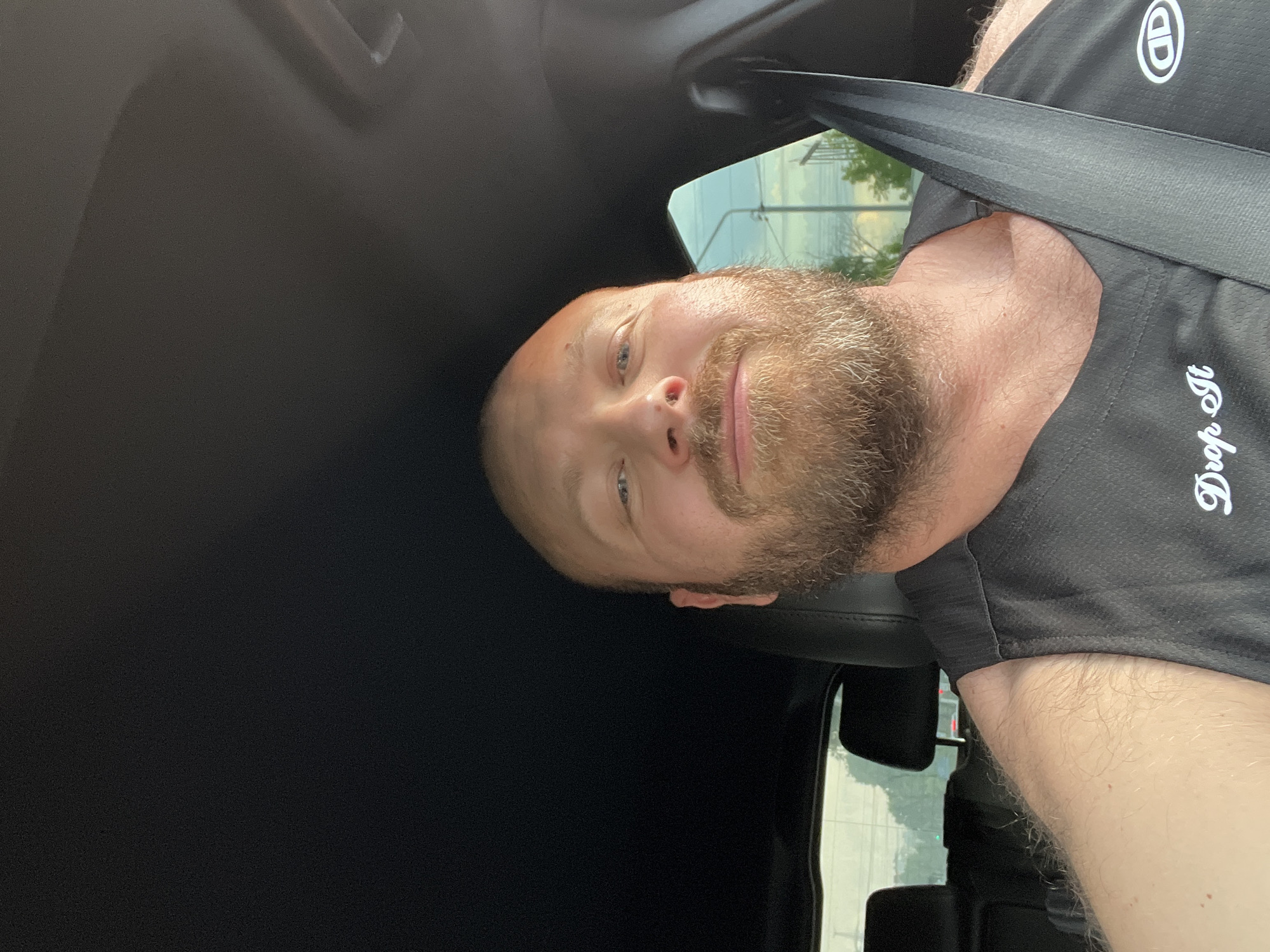 A man is pictured from the chest up, sitting in the driver's seat of a car. He is bald, with a light complexion and a full beard. He is wearing a black sleeveless shirt with white lettering that reads "Drop It" and a logo on the right side. A black seatbelt is diagonally across his chest. The interior of the car is visible, including the black headliner and part of the dashboard. Through the window, a cloudy sky and some greenery are visible. The location is identified as Chaikovsky, Russia.