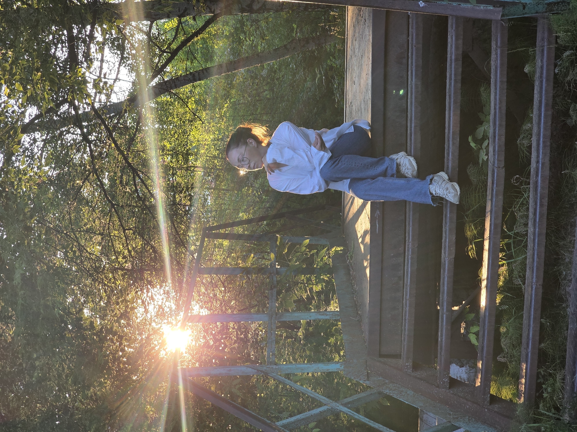 A young woman with glasses, wearing a white oversized shirt and blue jeans, sits on a metal staircase outdoors. She is looking to her left, with her right arm bent and her hand resting on her arm. The sun is setting, casting a warm glow through the trees and creating lens flares. The staircase appears to be old and possibly rusted, with a green metal railing. The surrounding environment is overgrown with greenery, suggesting a natural or semi-wild setting. The scene evokes a sense of peace and contemplation. The location is the city of Chaikovsky, Russia.