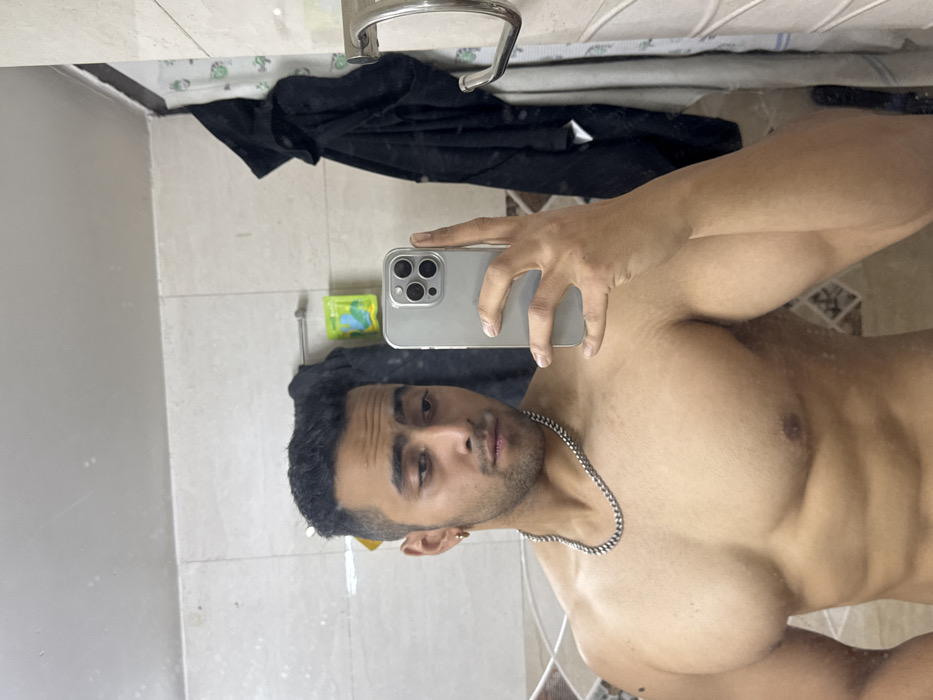 A man with dark hair and stubble is taking a mirror selfie in a bathroom. He is shirtless, wearing a silver chain around his neck. The background shows bathroom tiles, a towel rack with a black garment draped over it, and a soap dispenser. The man holds a smartphone with a gray case. The image is rotated counterclockwise.