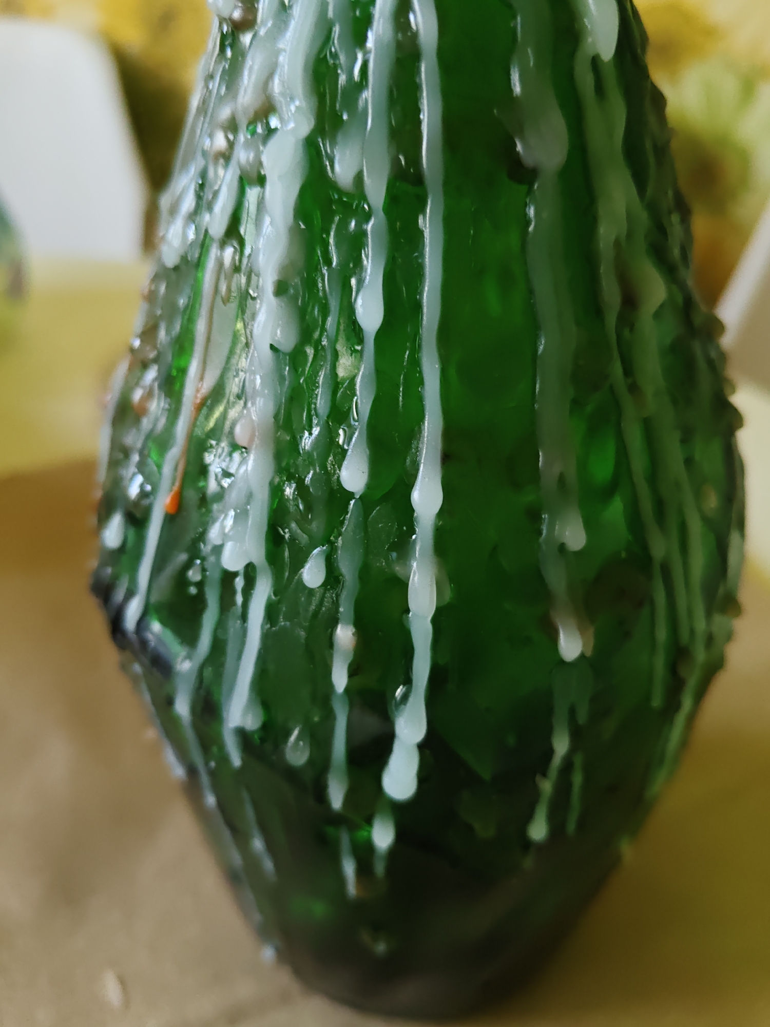 The image shows a close-up of a green glass bottle with white wax dripping down its sides. The bottle has an irregular, textured surface and appears to be partially obscured by the wax. The background is blurred, with areas of yellow and white suggesting an indoor setting. There are no people visible in the image, and no specific details about the city of Rostov-on-Don, Russia, can be inferred from the visual content. The lighting is soft, and the focus is on the bottle and the wax.