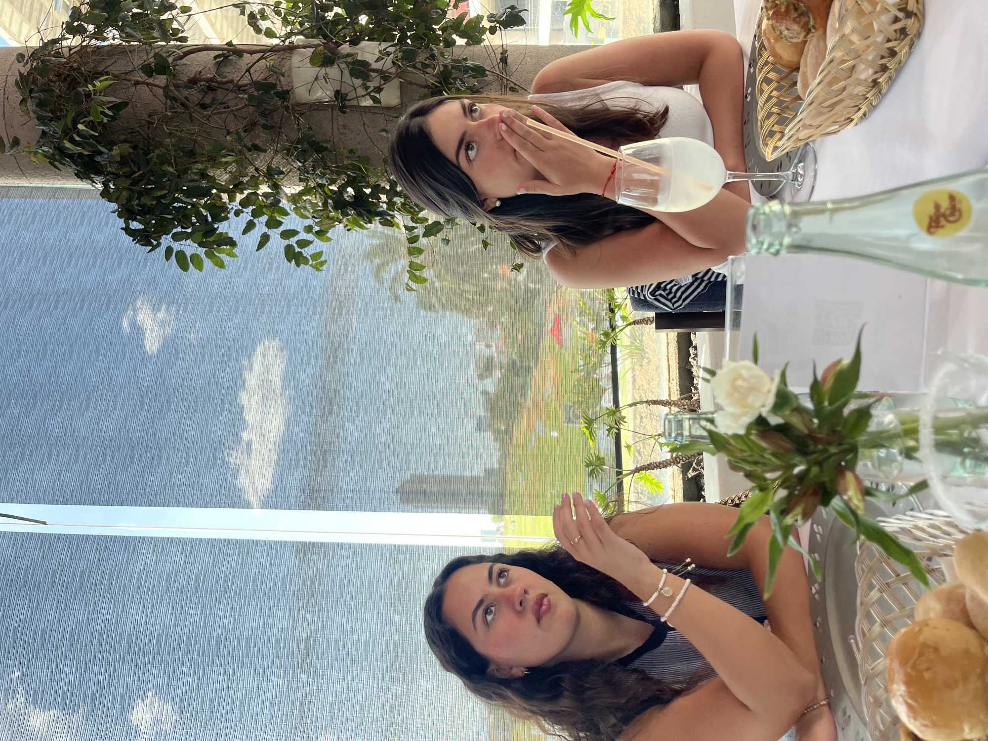 The scene depicts two young women seated at a table inside a restaurant or cafe in San Bernardino Tlaxcalancingo, Mexico. Both are positioned with a large window behind them, offering a view of an outdoor landscape.
The woman on the left has dark, curly hair and wears a striped top. She is looking upwards and to her right, with her right hand raised near her chin. She wears a gold ring on her right hand and two white beaded bracelets with a gold charm on her left wrist.
The woman on the right has long, dark hair and wears a white tank top. Her left hand is covering her mouth, with a light-colored straw positioned between her fingers and chin. She wears pearl earrings and a red string bracelet on her right wrist, and her gaze is directed upwards and to her left. A glass containing a light-colored beverage with a straw sits in front of her.
The table is covered with a white tablecloth. A small glass vase with white flowers and green foliage serves as a centerpiece. To the right, a wicker basket holds an assortment of bread rolls. A clear glass bottle, possibly of sparkling water, with a yellow label, is also on the table. A silver-colored charger plate is partially visible beneath a bread roll.
Beyond the window, a light blue sky with scattered white clouds is visible, partially obscured by horizontal blinds or shades. A lush green landscape, possibly a golf course, stretches into the distance, with a tall, modern building visible on the horizon. Palm trees and other vegetation are present outside the building. Inside, a concrete column covered in climbing green vines stands to the right of the frame, near the window.