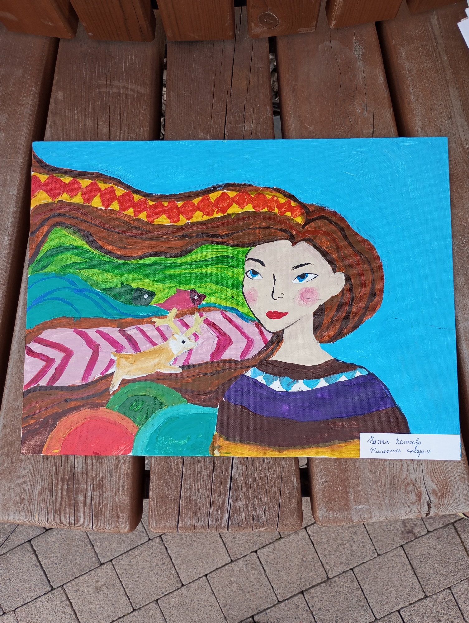 The image shows a painting resting on a wooden surface that appears to be part of a picnic table. The table itself is outdoors, evidenced by the brick ground visible in the lower portion of the image. The painting depicts a stylized portrait of a woman with light skin, bright blue eyes, and red lips. Her hair is brown and extends upward into a colorful pattern featuring red, yellow, brown, green, and blue hues. A small deer-like creature is also depicted within the colorful section of the painting. The woman is wearing a garment with brown, purple, and blue stripes, and the background of the painting is solid blue. A small white label bearing text in Russian is affixed to the lower right corner of the painting. The text likely contains the artist's name and information. The scene is situated somewhere in Pervomaysky, Russia, as per the instructions.