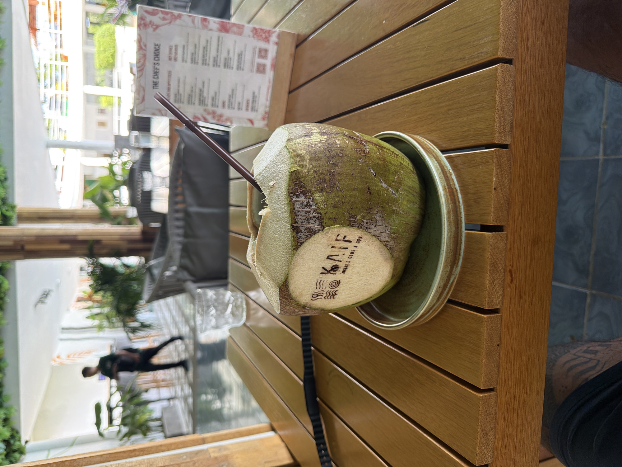 The media file captures a close-up, angled view of a fresh coconut drink resting on a light-brown slatted wooden table, likely in an outdoor or semi-outdoor dining area. The coconut, primarily green with some white husk showing where it has been opened, sits in a green ceramic saucer and has a dark brown straw inserted into it. A visible logo carved into the coconut reads "KAIF" with smaller text underneath saying "BEACH CLUB & SPA", accompanied by several stylized wave or line designs.
In the upper left portion of the image, a menu stands upright on the table. Although largely out of focus, the heading "THE CHEF'S CHOICE" is clearly visible, along with sections labeled "HOT DISHES" and "MAIN". The background, significantly blurred, reveals bright natural light, green foliage, and what appears to be part of a building or structure. A distinct, blurry silhouette of a person, possibly male, is seen walking in the distant background.
The setting suggests a warm, daytime environment, consistent with the location context of Kathu, Thailand, and the presence of a coconut drink. The overall impression is one of leisure and relaxation. A black strap, possibly from a bag or camera, lies on the table near the coconut. On the far right, a dark-clothed leg with visible tattoos can be partially seen, suggesting the perspective of someone seated at the table.