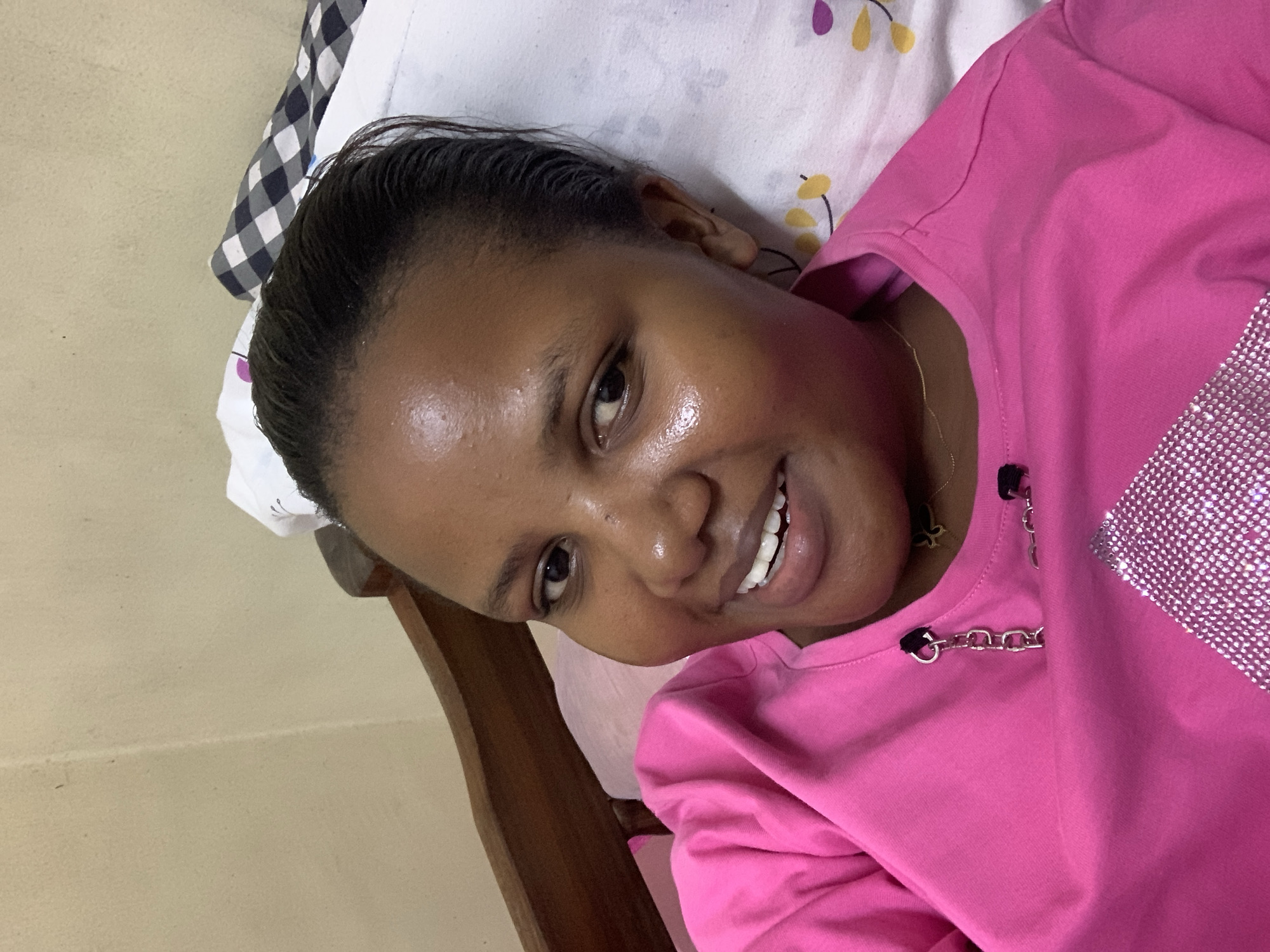 A close-up shot shows a woman with dark hair pulled back, lying on a pillow and smiling at the camera. She is wearing a bright pink shirt with rhinestone embellishments and a silver chain detail at the neckline. The pillow has a white background with a pattern of yellow and purple floral shapes. The background also includes a wooden bed frame and a light-colored wall. There is no information to determine that the location is in the city of Dar es-Salaam, Tanzania.