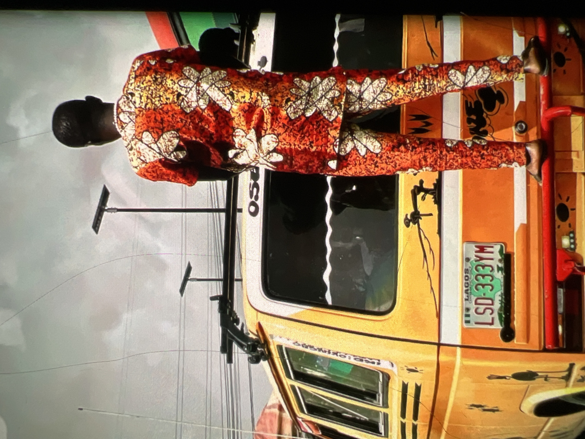 A rear view of a person standing on the back bumper of a yellow van in Lagos, Nigeria. The person is wearing an orange, red, and white patterned shirt and pants, and dark brown shoes. The van has a white stripe across the back and a license plate that reads "LSD-333YM LAGOS". The sky is overcast and there are power lines and streetlights visible in the background. There are also several dark, silhouetted figures in the background. The van appears to be a commercial transport vehicle, possibly a "danfo" bus, common in Lagos.