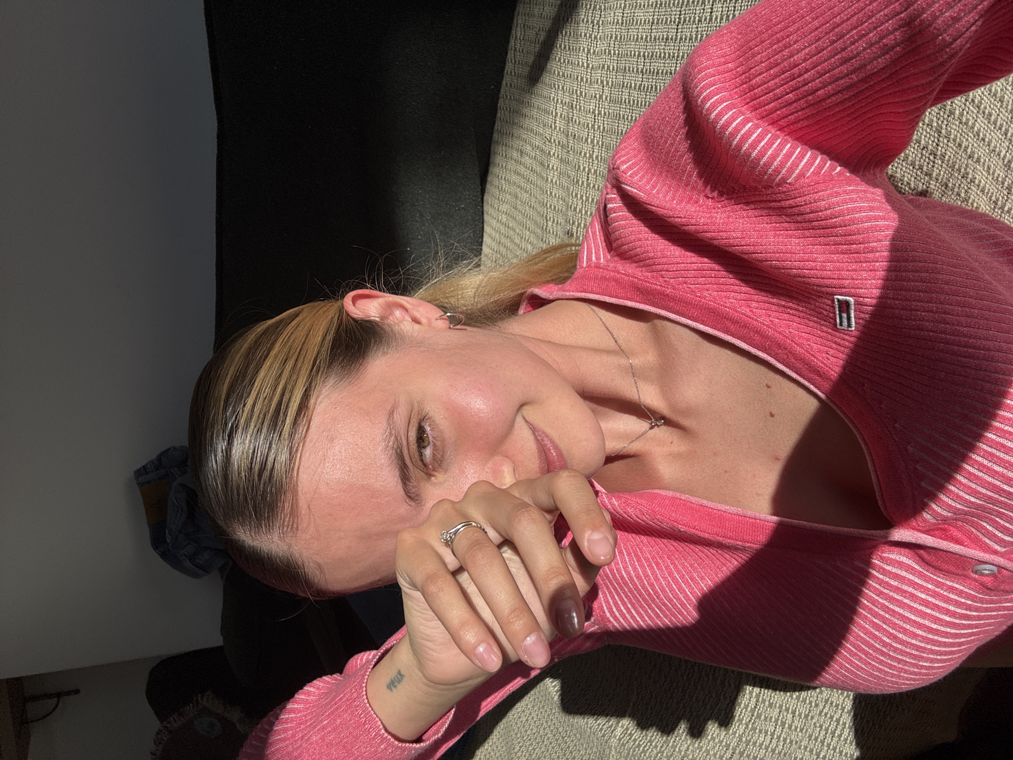 A young woman with blonde hair, pulled back from her face, is depicted in a close-up shot, looking towards the camera with a gentle expression and a slight smile. She is lying down, resting her head on a light-colored, textured surface, likely an upholstered sofa or chair.
She is wearing a bright pink, ribbed long-sleeved top or cardigan featuring thin white horizontal stripes. A small rectangular logo, which appears to be red, white, and blue, is visible on the left chest area of her top. She accessorizes with a silver ring on the fourth finger of her right hand, a small silver hoop earring in her left ear, and a delicate silver necklace with a tiny pendant around her neck. A small tattoo reading "veux" is visible on her left wrist.
Her right hand is raised, partially covering her mouth and chin, a gesture that could convey shyness or contemplation. Strong, direct sunlight streams into the indoor setting, casting sharp shadows across her face, hair, clothing, and the surrounding surfaces. This suggests it is daytime, likely morning or late afternoon. The background includes a dark, unidentifiable surface behind her head and the textured fabric where she rests.
Notable details include a visibly discolored or bruised nail on her ring finger of her right hand, and small red marks visible on her décolletage. The overall mood is relaxed and natural, highlighted by the natural lighting.