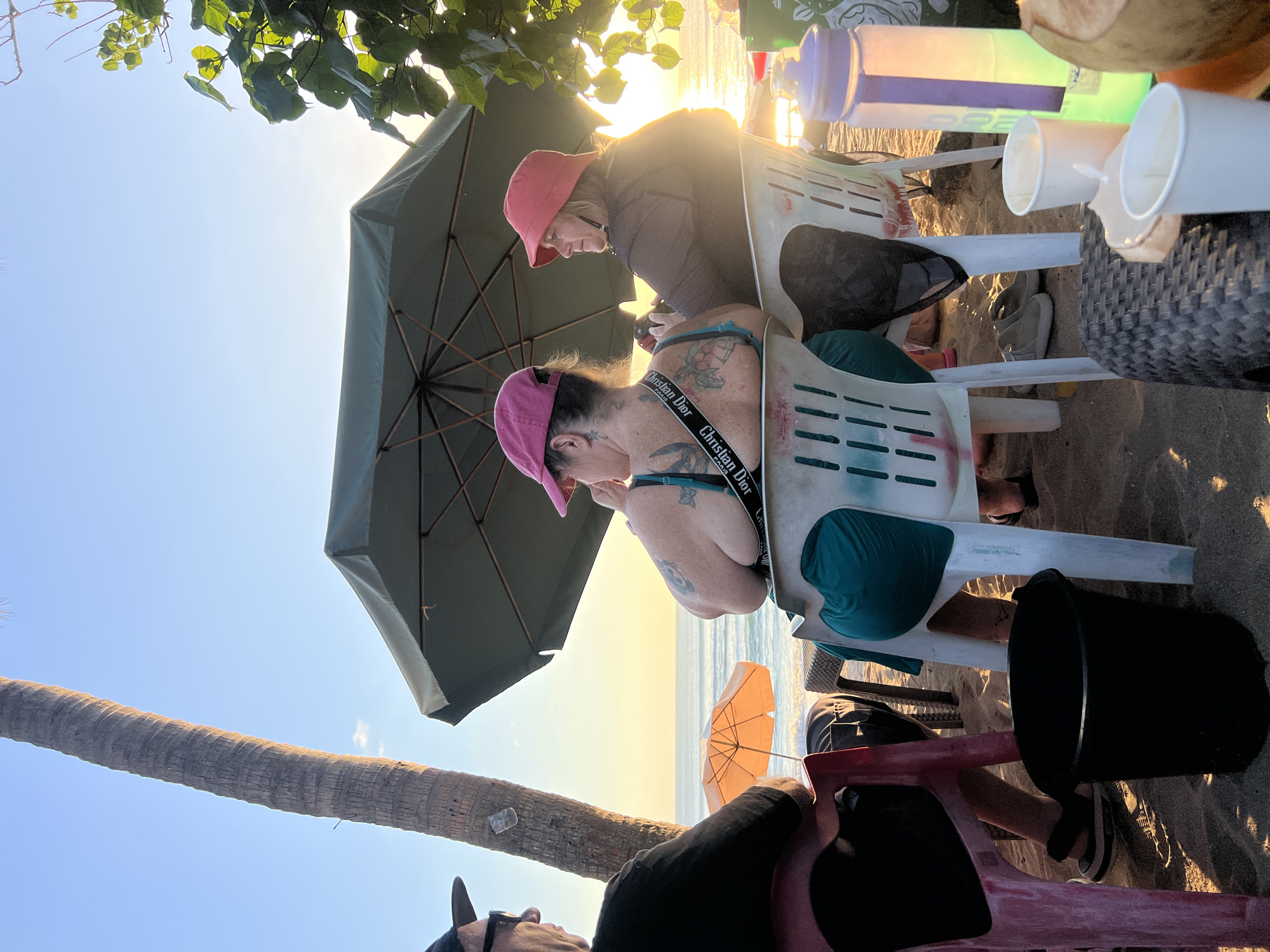 The image depicts a relaxed scene on a beach in Kuta, Indonesia, during sunset. Two women are seated in plastic chairs under a large green umbrella, facing the ocean. The woman on the left is wearing a pink baseball cap and a teal swimsuit, with tattoos visible on her back and shoulders. The woman on the right wears a red bucket hat and a brown top. A third person, a man wearing a dark cap and sunglasses, is partially visible on the far left. In the foreground, a black bucket sits on the sand, and a woven table holds two paper cups and a water bottle. A coconut is also visible on the table. The background features the ocean with gentle waves and the golden glow of the setting sun, framed by palm trees and lush foliage. The atmosphere is tranquil, suggesting a leisurely end to the day.