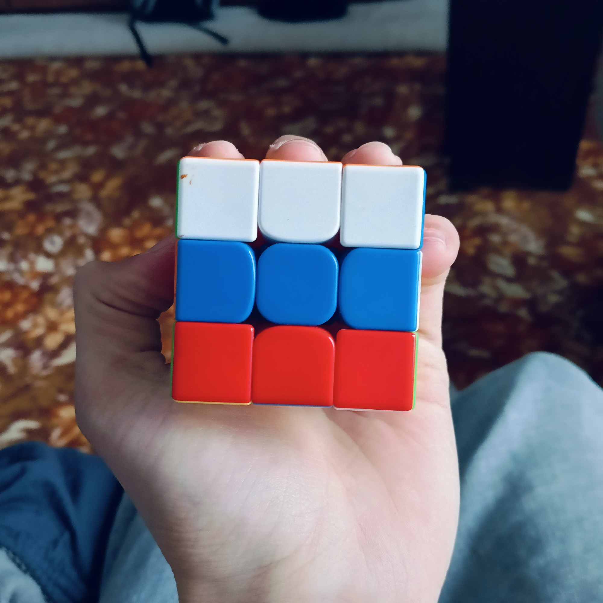 A hand holds a Rubik's cube displaying a face with white, blue, and red squares, reminiscent of the Russian flag. The background is blurry, showing part of a chair and patterned flooring. The location in the city of Буренка, Russia, cannot be determined from the image.