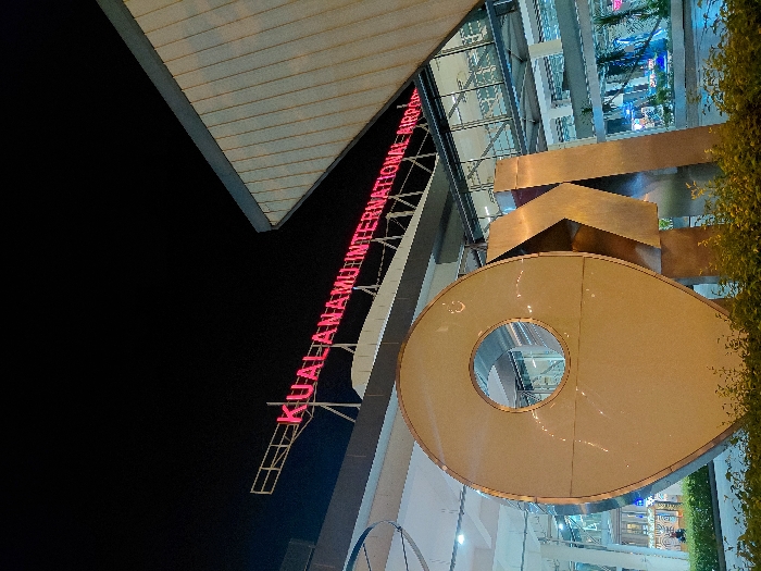 The image depicts the exterior of Kualanamu International Airport, situated in the city of Sidourip, Indonesia, at night. In the foreground, a large, metallic, three-dimensional sculpture resembling a geographical pin icon, with a circular cutout, stands amidst low green foliage. To its right, partially visible, are blocky metallic letters appearing to form "KNO." Behind this sculpture, the modern airport building is illuminated, featuring large glass facades and an elevated glass-enclosed walkway. Mounted high on the building's structure, a prominent red neon sign displays the text "KUALANAMU INTERNATIONAL AIRPORT." The sky above is dark, indicating nighttime, with artificial lighting from the airport structure and the neon sign providing illumination to the scene. No discernible individuals or specific activities are visible in the immediate foreground, though distant interior lights suggest activity within the airport building.