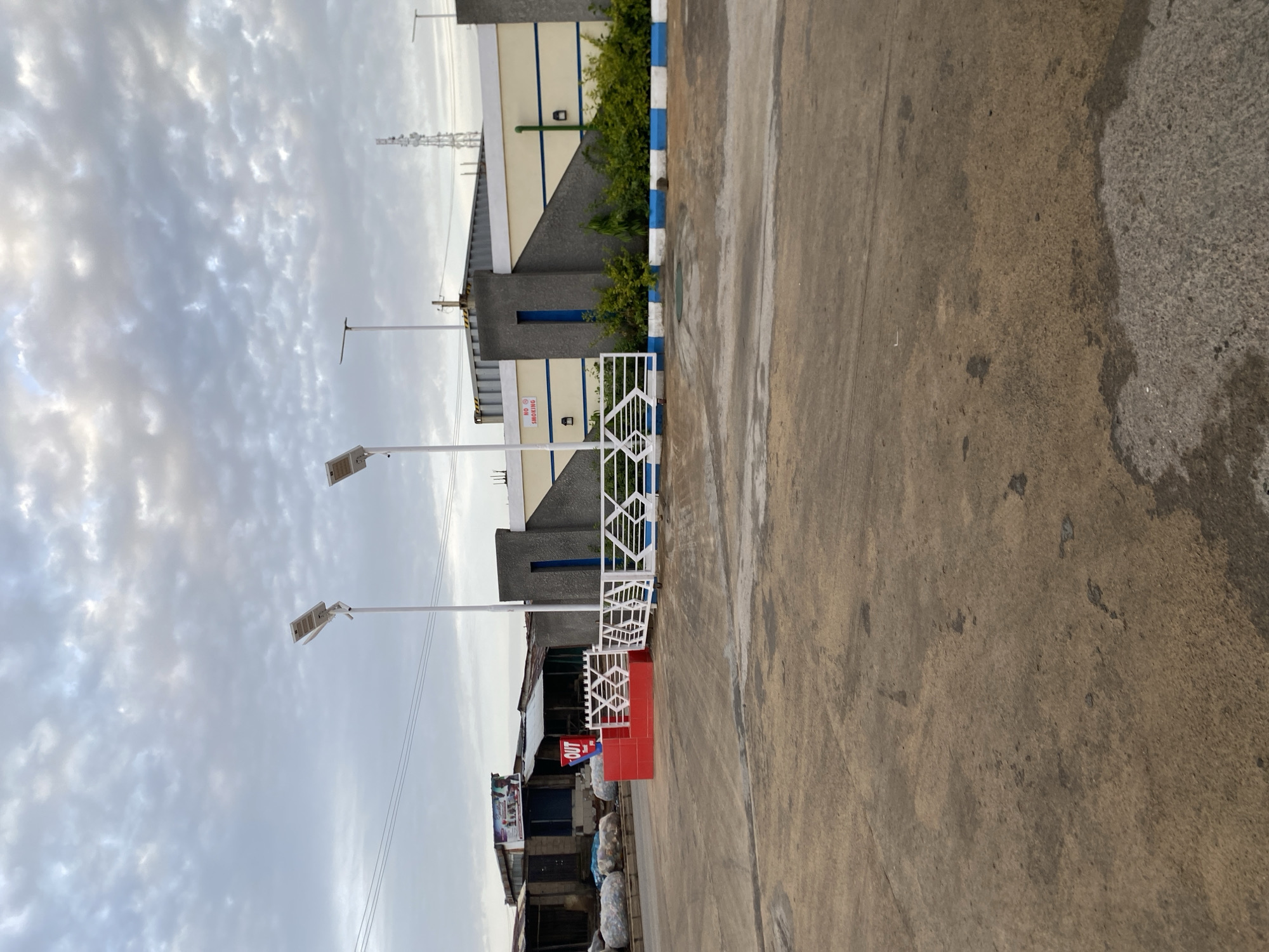 The image depicts an outdoor scene on a cloudy day, likely in Barakin Lerawi, Nigeria. In the foreground, a paved area with patches of wear and tear leads towards a gate. The gate itself is white with a patterned metal design, flanked by a red barrier on the left with the word "OUT" in blue lettering. Behind the gate and barrier is a paved area with some vegetation. Further back, a building complex is visible, featuring a wall with a yellow and blue design, a blue vertical accent, and decorative grey pillars. Two streetlights are prominently positioned in the mid-ground, extending from white poles. To the left of the gate, a row of informal structures with corrugated metal roofs are present, with piles of what appear to be plastic bags or bales of waste material in front of them. A tall telecommunications tower can be seen in the distance against the sky. The overall impression is of an industrial or commercial area with evidence of waste management activities.