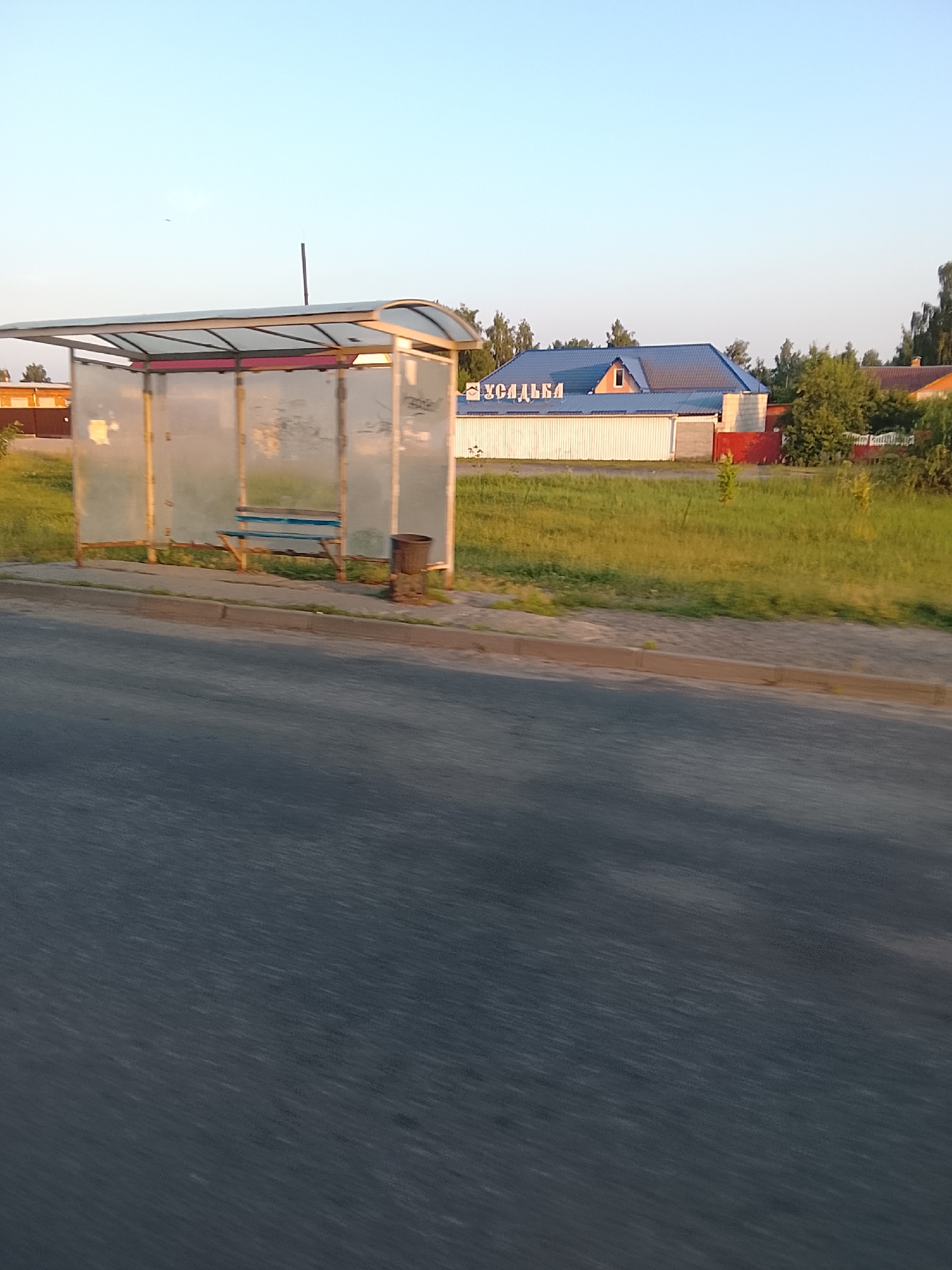 The image shows a bus stop shelter on the side of a road in Zhlobin, Belarus. The shelter is made of metal and has glass panels that are partially obscured by graffiti. A blue bench is visible inside the shelter. A trash can is located next to the shelter. In the background, there is a building with a blue roof and the word "УСАДЬБА" written on it in blue letters. The area around the bus stop is grassy, and there are trees in the distance. The sky is clear and blue, suggesting it is daytime. The road in the foreground is asphalt and appears to be wet.