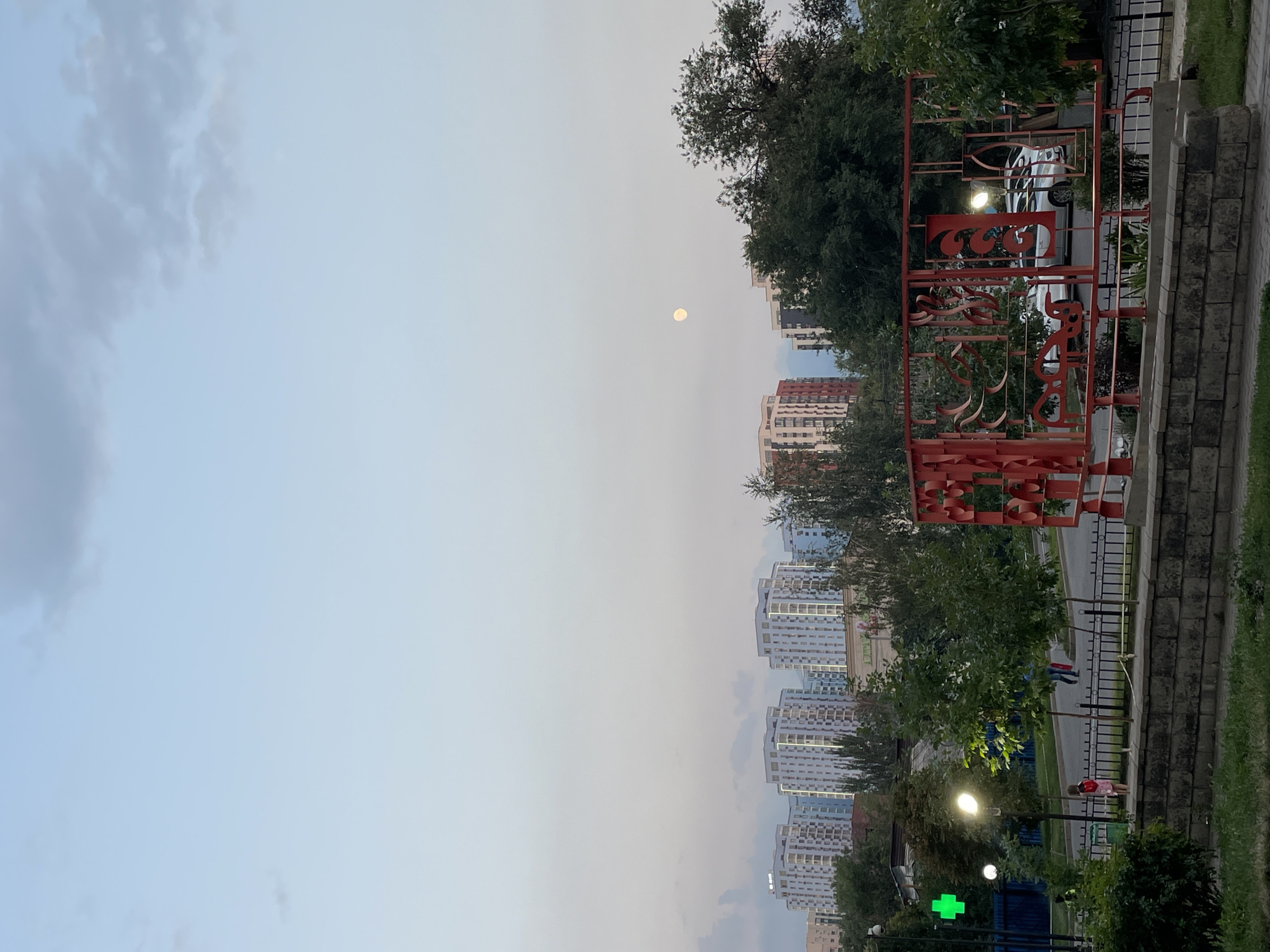 The image depicts an evening scene in the city of Almaty, Kazakhstan, likely in a residential area. In the background, a row of modern apartment buildings with illuminated windows are visible against a pale sky with scattered clouds. A full moon is present in the upper right quadrant of the sky. In the foreground, a decorative red metal structure is prominent, featuring an intricate, abstract design. Through the structure, a street is visible with parked cars and some streetlights. To the left, there is a green pharmacy cross sign, a lamppost with a bright light, and several trees. A few individuals are seen walking on the sidewalk or street, including a child wearing a red dress. The overall environment suggests a mixed-use urban setting with residential and commercial elements.