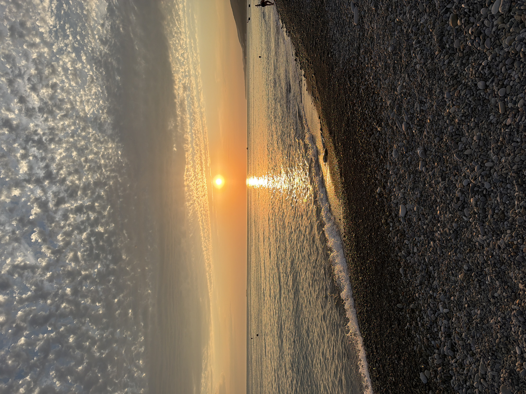 The image depicts a sunset over the Black Sea from a pebble beach, likely located in Sochi, Russia. The sun, a bright orange orb, is low on the horizon, casting a golden reflection across the calm water. The sky is filled with scattered, fluffy clouds, illuminated by the setting sun. The foreground is dominated by a textured expanse of grey and brown pebbles that make up the beach. Small waves gently break on the shore. In the distance, a faint coastline is visible with a hilly landscape. A few dark buoys are present in the water. To the right of the frame, a solitary person is visible on the beach, their posture suggesting they are facing the sea.