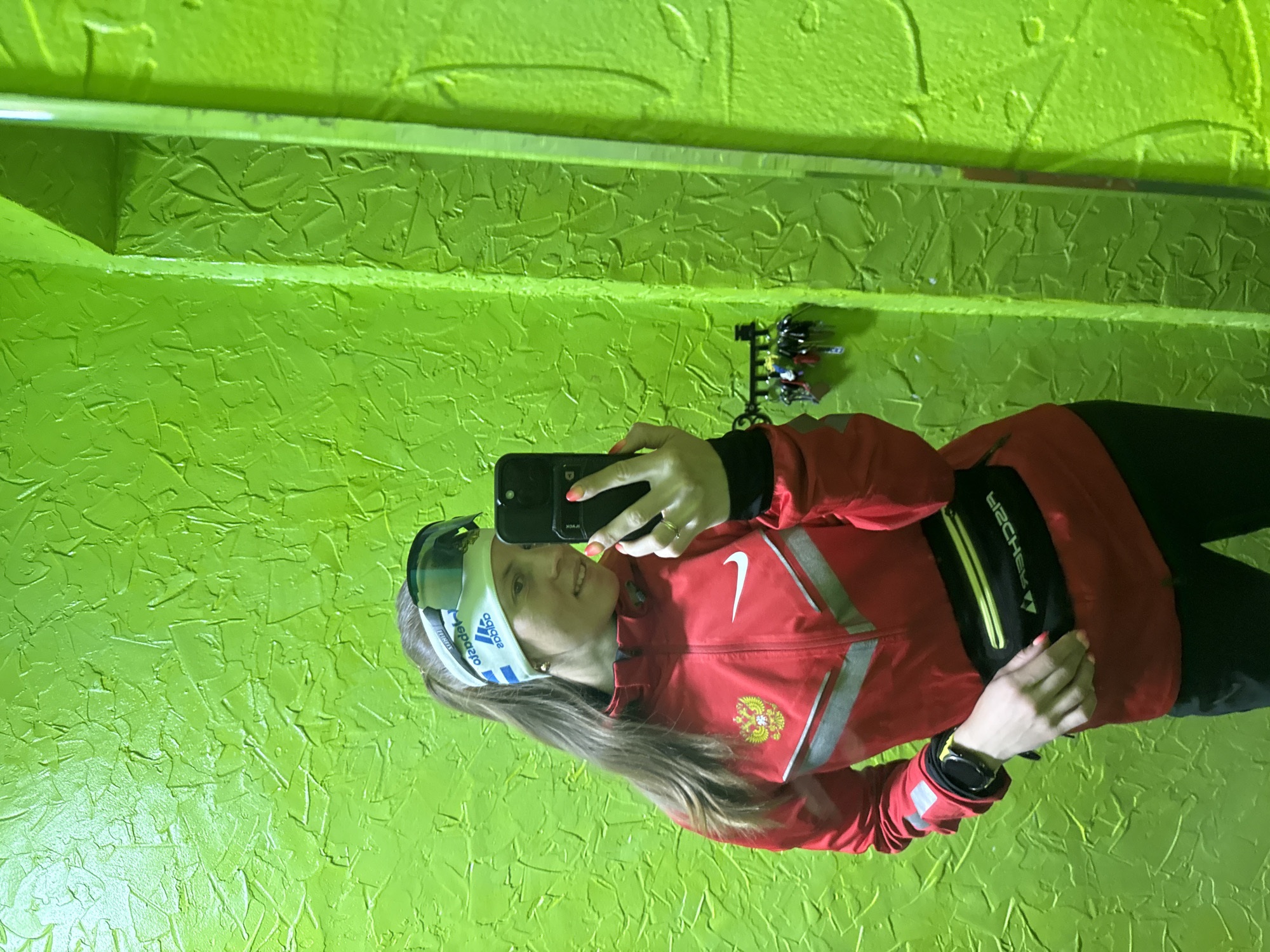 A young woman is taking a selfie in front of a green textured wall, likely in Chaikovsky, Russia. She wears a red Nike jacket with the Russian coat of arms, a black fanny pack, black leggings, and a headband with ski goggles. She holds a black smartphone to take the picture. Keys hang on the wall behind her. She also wears a watch on her left wrist.