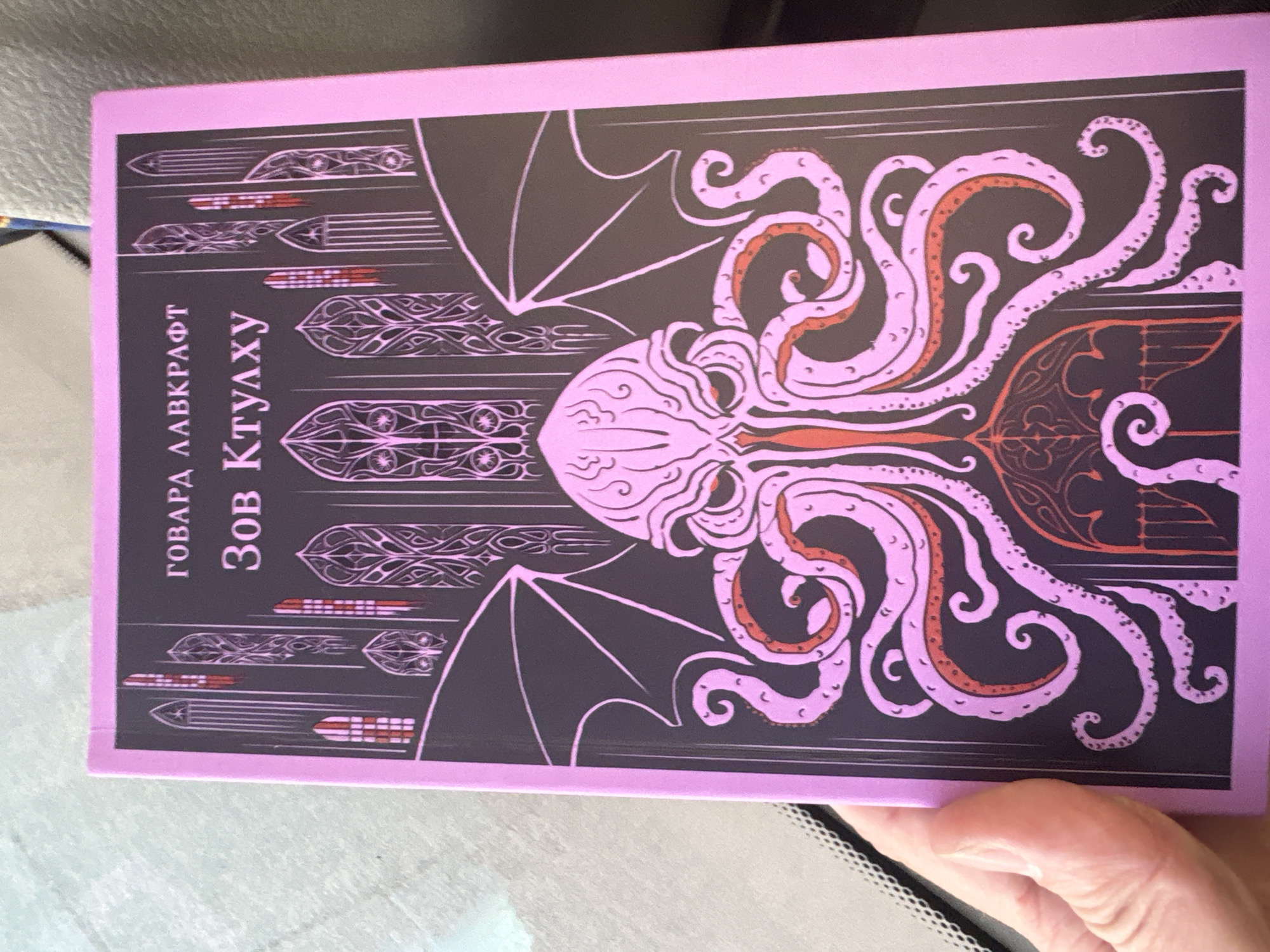 The image shows a copy of H.P. Lovecraft's "The Call of Cthulhu" (Зов Ктулху) in Russian. The cover features a stylized depiction of Cthulhu. There is no evidence in the image to suggest the location is Saint Petersburg, Russia. The image appears to be taken indoors. A person's hand is partially visible.