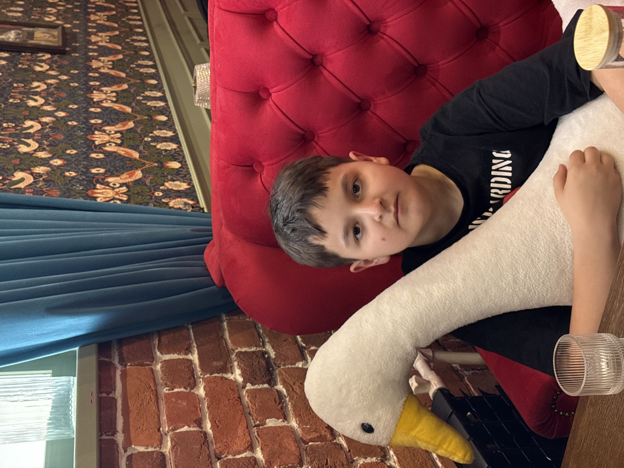 A child is lying on a red tufted chair, hugging a large, white plush goose. The child has short brown hair and is wearing a black t-shirt with white lettering. A glass with a wooden lid is visible near the child's head, and another clear, ribbed glass is visible near the goose's tail. A brick wall is in the background, along with a blue curtain or drape. A patterned wallpaper is also visible. The location is Nizhnevartovsk, Russia.