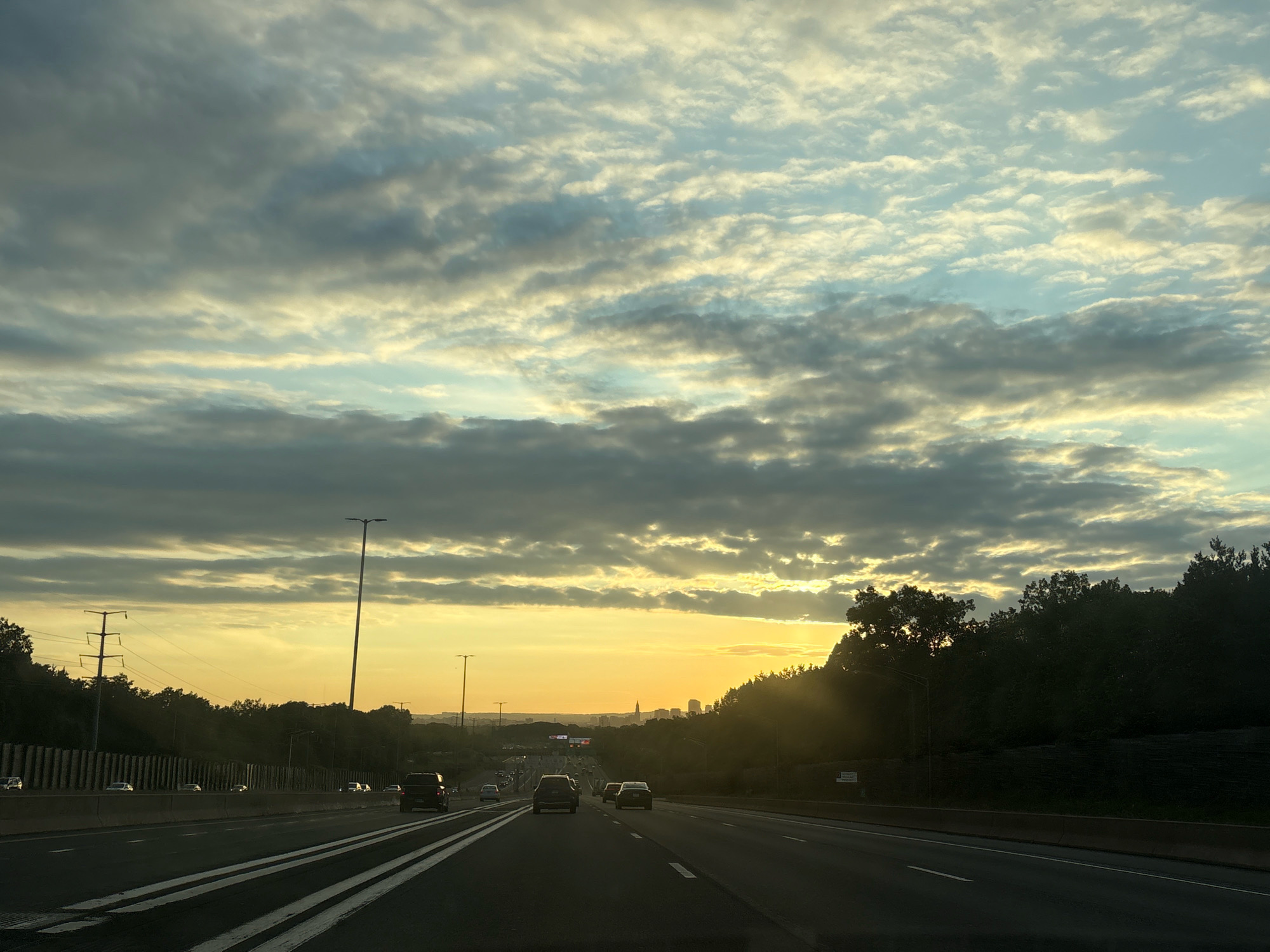 A wide shot captures a highway at dusk with a city skyline in the distance, likely East Hartford, Connecticut, USA. The sky is filled with dramatic clouds, tinged with the warm hues of the setting sun which filters through gaps in the cloud cover. Multiple lanes of a highway are visible, with cars driving in both directions. The road is well-lit by streetlights, and guardrails line the sides. Trees and foliage form a dark silhouette against the bright sky on the right side of the frame. In the far distance, the shapes of buildings suggest a metropolitan area. The image conveys a sense of travel and the transition from day to night.