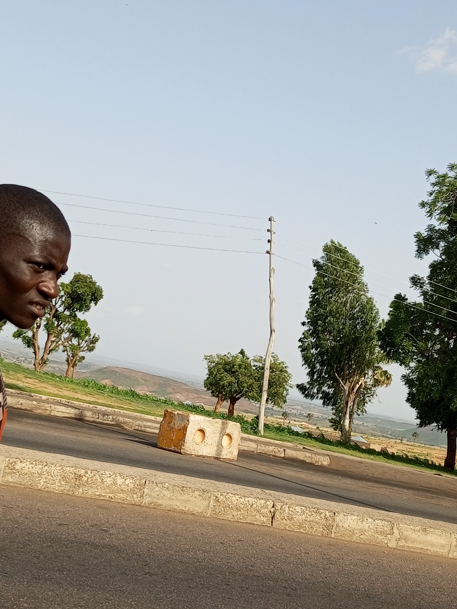 The image captures a street scene in Gombe, Nigeria. In the foreground, a young Black male is partially visible on the left, looking towards the right with a stern expression. He is wearing a dark shirt with an orange and white striped pattern. The street, paved with asphalt, curves towards the right. A concrete curb, detailed with textured stones, lines the edge of the road.
Further down the road, a concrete barrier block, painted yellow and white, is situated on the side of the street. Beyond the road, a grassy embankment slopes upwards, dotted with several trees. In the background, a panoramic view of the Gombe landscape unfolds, characterized by rolling hills and sparse vegetation. Power lines stretch across the frame, supported by wooden utility poles. The sky is a pale blue, with a few wispy clouds. The overall impression is of a typical day in a semi-urban or rural area of Gombe, Nigeria.