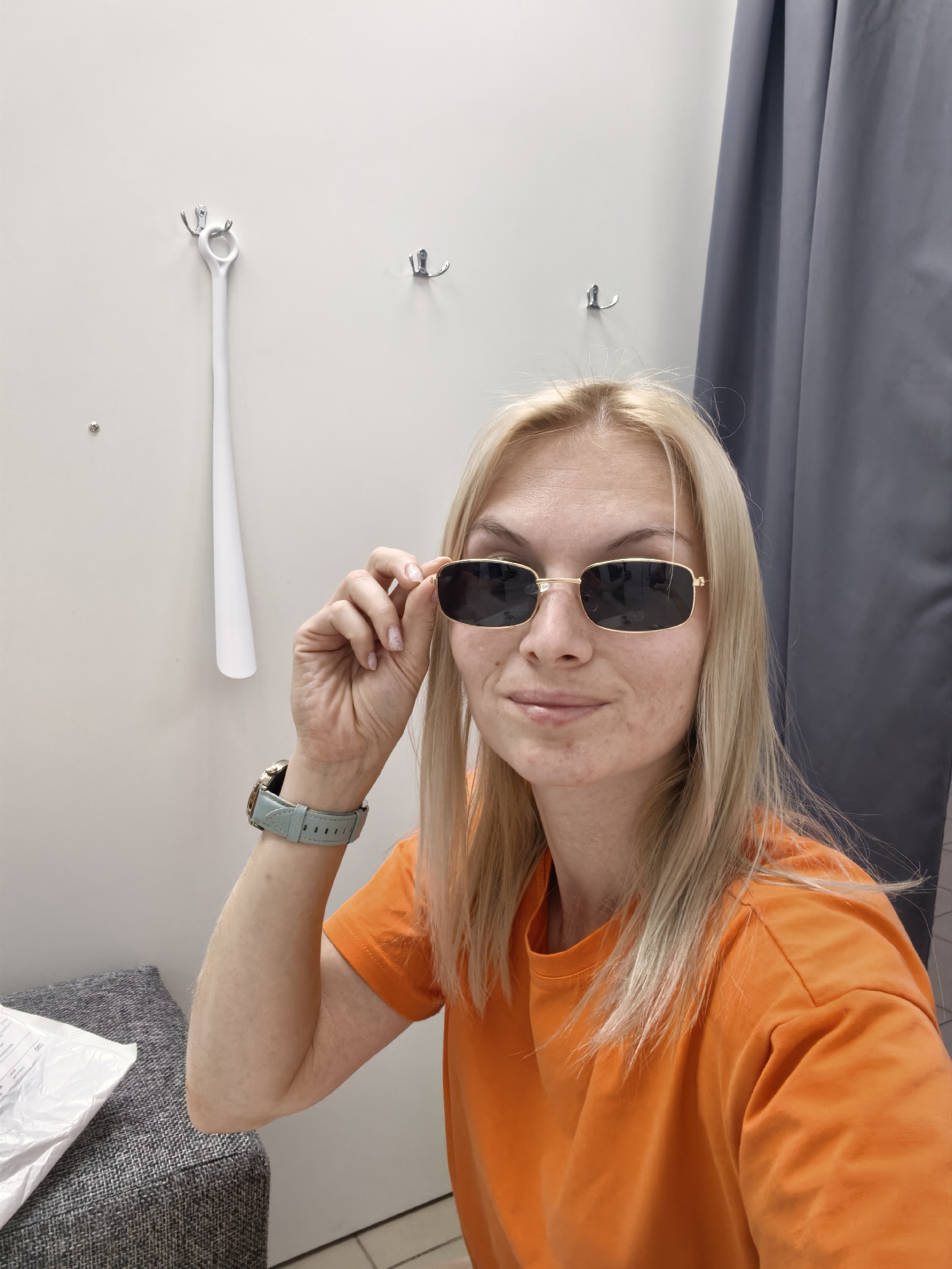 In an indoor fitting room, a blonde woman in an orange t-shirt and sunglasses is taking a selfie. She is adjusting her sunglasses with her right hand, and her left arm is resting on a grey upholstered bench. A pale blue watch is visible on her left wrist. Behind her, a white shoehorn hangs on a hook attached to a light-colored wall. A gray curtain is visible to her right. The image appears to be taken in Sterlitamak, Russia, but this cannot be confirmed from the image alone.