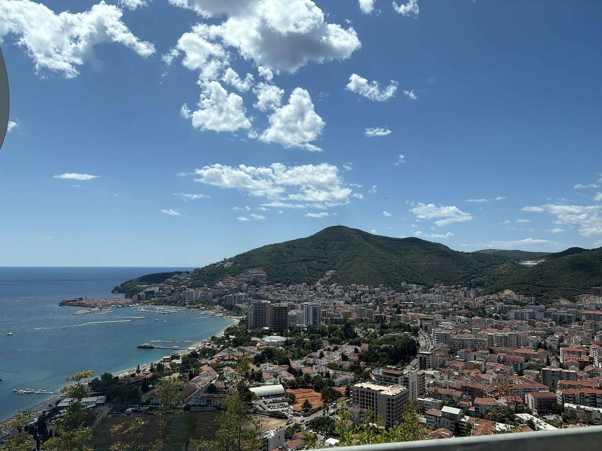 This is a high-angle, full shot of a coastal city in Košljun, Montenegro, on a clear, sunny day. The scene is dominated by a large, green, mountainous hill that forms the backdrop to the city. The city itself is a dense collection of buildings with red-tiled roofs, interspersed with taller, modern structures. The coastline is visible on the left, with the blue ocean meeting a shore dotted with buildings and a marina filled with boats. A long pier extends into the water from the city. In the foreground, there are some trees and a metal railing, suggesting the vantage point is from an elevated observation area. The sky is a bright blue with scattered white clouds. There are no people visible in the image.