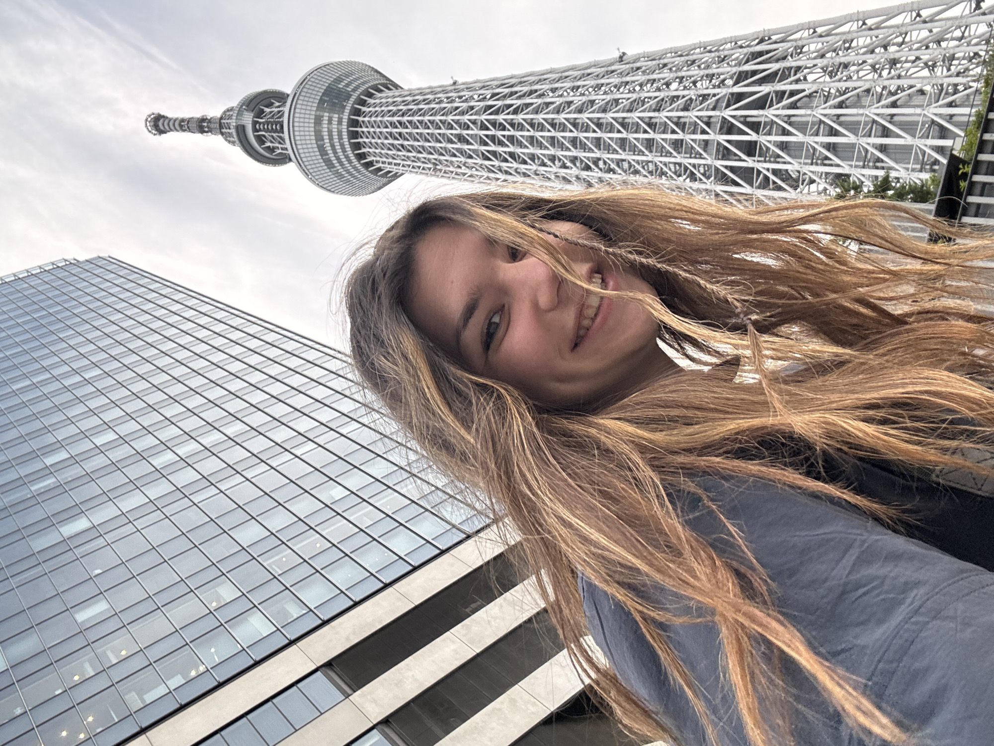 A low-angle, full-body selfie shows a young woman in the foreground smiling at the camera, her face partially obscured by her long, wavy, light brown hair. She is wearing a dark blue jacket. Behind her, to the left, is a modern glass-fronted skyscraper, and to the right, the Tokyo Skytree, a tall, lattice-like observation and broadcasting tower. The sky is overcast. The image appears to have been taken outdoors in the city of Sumida, Japan, given the presence of the Tokyo Skytree.