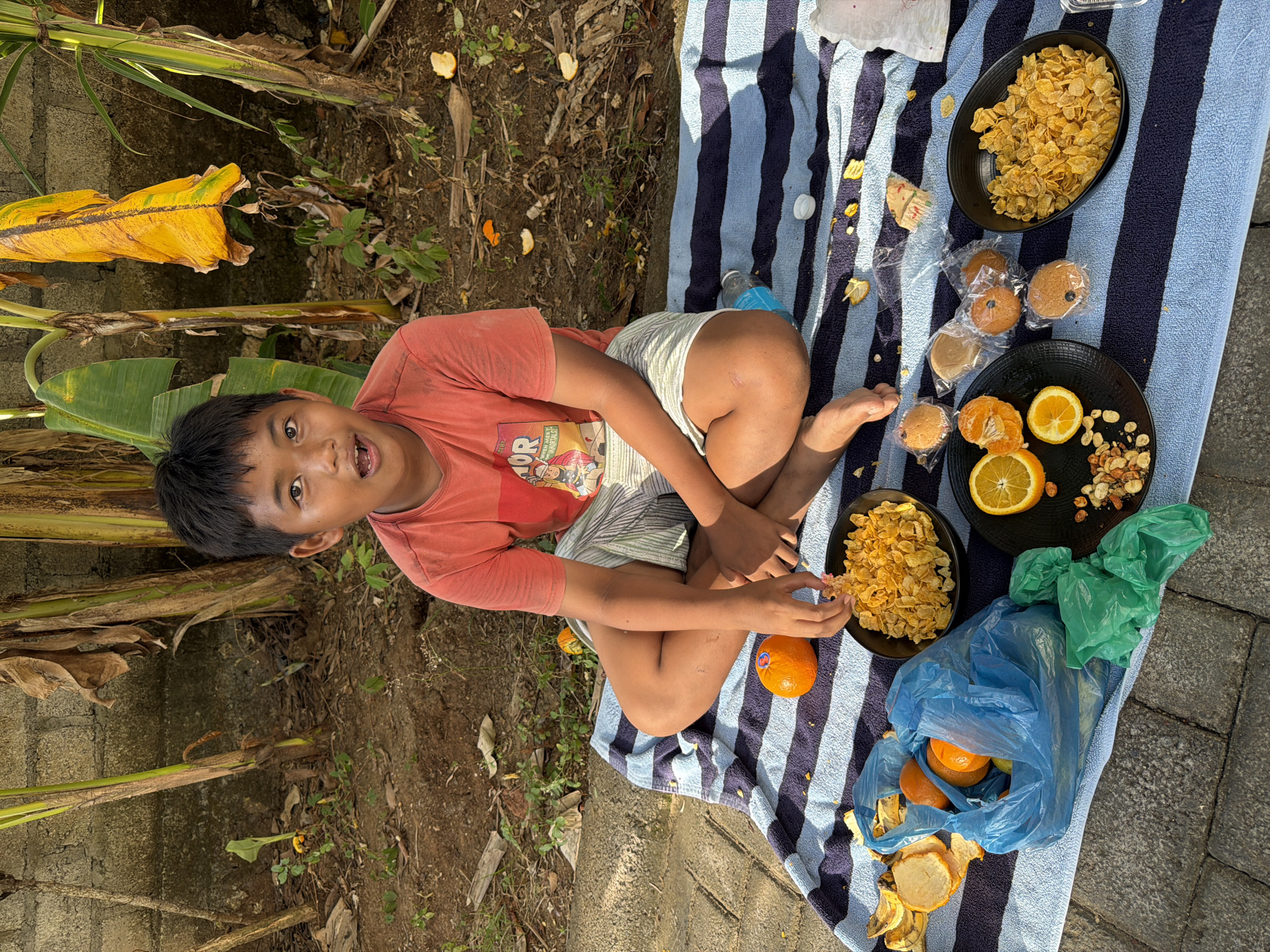 In Kuta Selatan, Indonesia, a young boy sits cross-legged on a striped blue and white blanket outdoors, with a wall and banana trees in the background. The boy, wearing an orange t-shirt and striped shorts, has an open mouth, appearing to be speaking or reacting to something. In front of him are two black bowls, one filled with cornflakes and the other with sliced oranges, nuts, and some cornflakes. A blue plastic bag contains oranges and banana peels, with green plastic bags nearby. Several small, individually wrapped baked goods, possibly muffins or cookies, are also visible. Scattered around the area are pieces of orange peel, consistent with the fruit being eaten. The ground is dirt and stone, indicating an outdoor, possibly less-maintained area.