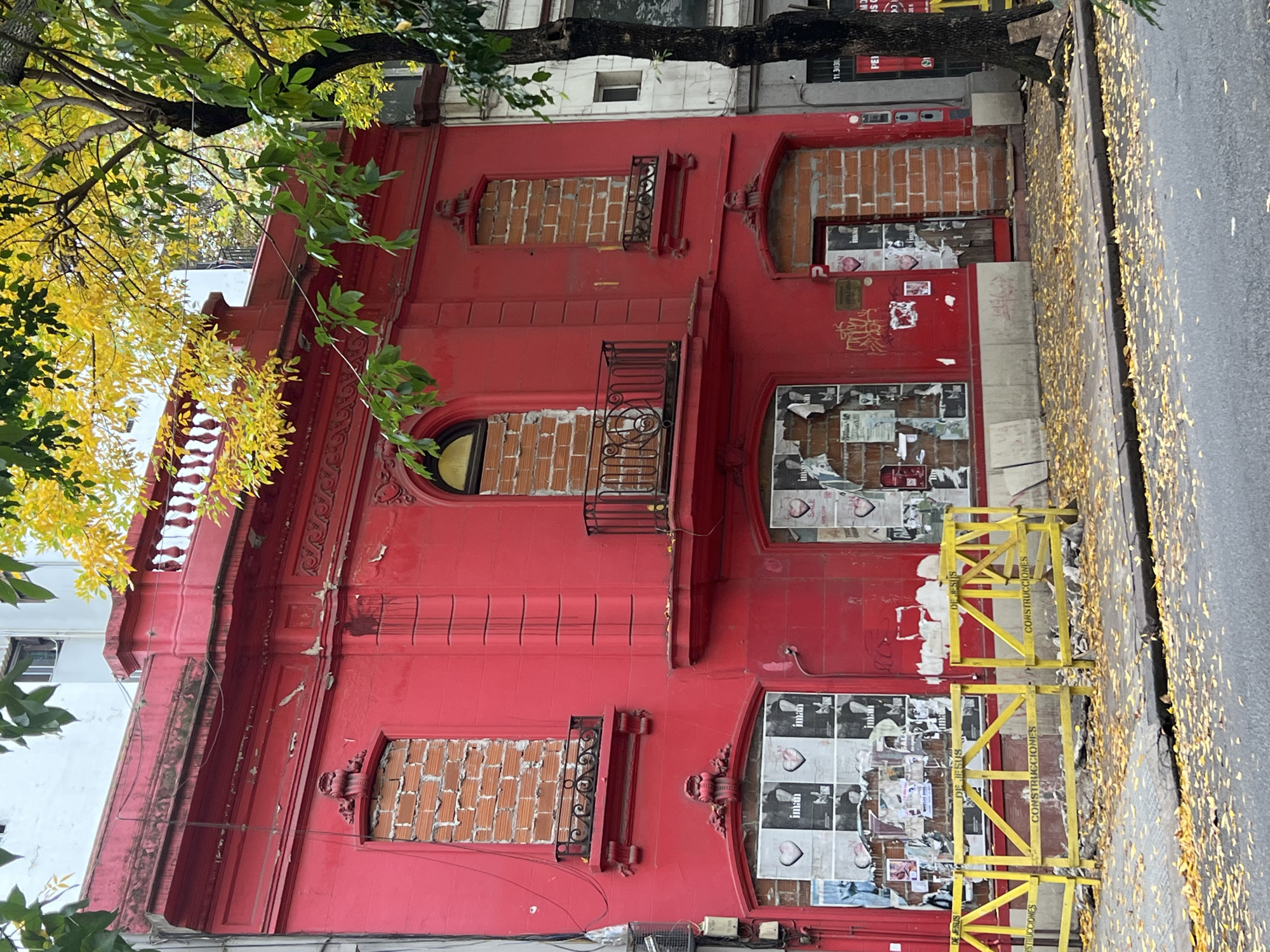 A three-story, red-painted building stands on a street in Buenos Aires, Argentina. Windows on the upper floors are bricked over, while those on the lower floors are covered with posters and graffiti. A yellow construction barrier sits in front of the building, and fallen leaves litter the sidewalk. Deciduous trees with yellowing leaves frame the upper left corner of the shot.