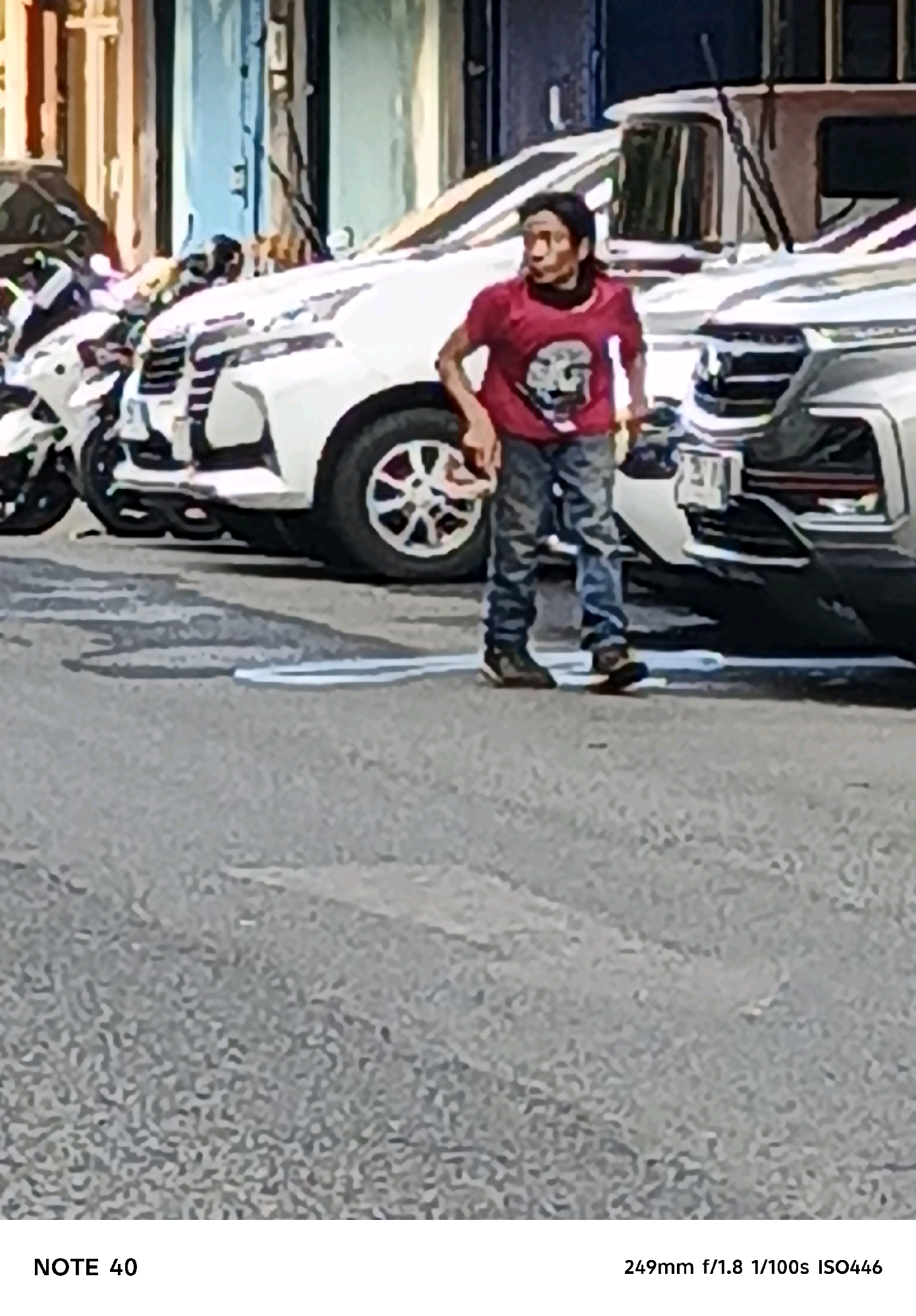 A young male, appearing to be in his late teens or early twenties, stands on an asphalt street in North Jakarta, Indonesia, wearing a red t-shirt with a graphic print and camouflage pants. He is looking towards the left of the frame with a surprised or concerned expression. His posture suggests he may have just been startled or is observing something of interest. The street is lined with parked vehicles, including a white SUV and motorcycles, indicating a busy urban environment. The background shows buildings with a mix of colors and architectural styles, common in Indonesian cities. The lighting suggests it is daytime, possibly late afternoon, with shadows cast on the street. The overall scene depicts a candid moment of an individual amidst the hustle and bustle of a city street.