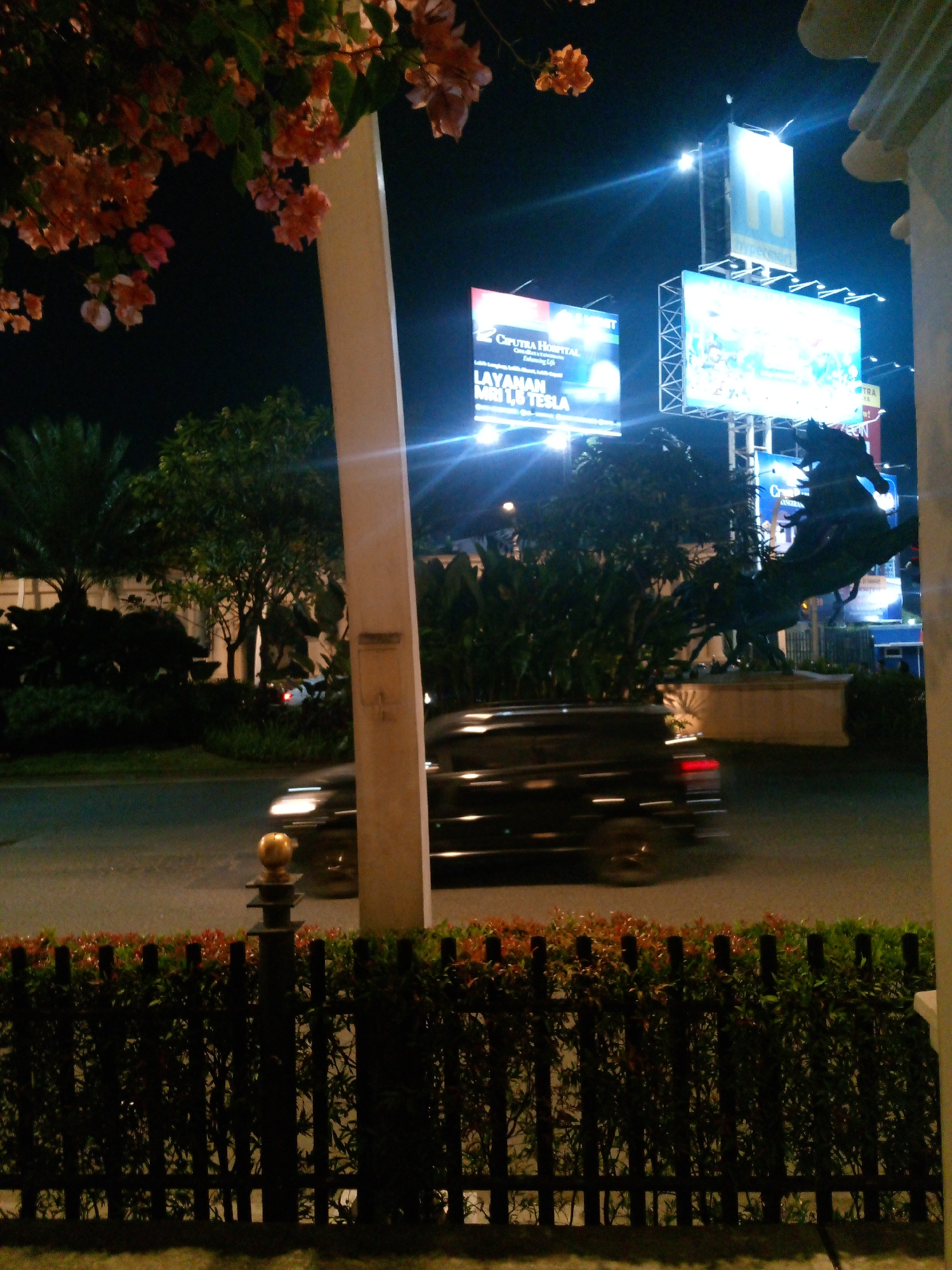 The image captures a nighttime scene in Bitung Jaya, Indonesia, featuring a blurred black car in motion on a road. The car's motion blur suggests a moderate speed. To the left of the road, a dark green hedge with reddish-brown foliage is visible, separated from the road by a black metal fence with vertical posts. A dark-colored metal fence post with a layered top, topped with a small golden sphere, stands in the foreground. Behind the hedge and fence, there's a landscape with trees and foliage. In the background, several illuminated billboards are prominent. One billboard on the left displays "CIPUTRA HOSPITAL" and "LAYANAN MRI 1,5 TESLA". Another large billboard in the center is brightly lit with blue and white lights. A tall, slender white pillar is situated to the right of the frame, partially obscuring the view of the billboards. Overhead, branches with small orange and pink flowers hang into the upper left corner of the image. The overall environment appears to be an urban or semi-urban setting at night, illuminated by artificial lights from the billboards and streetlights.