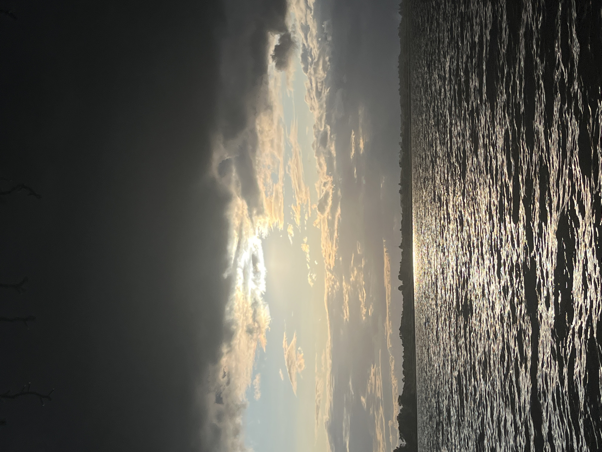 A dramatic sunset casts shimmering light across a choppy body of water, likely a river or lake, near Radchenko, Russia. Dark, heavy clouds dominate the upper portion of the frame, with a sliver of the sun breaking through near the center, illuminating the sky with golden and pale blue hues. The horizon line is defined by a dark silhouette of trees and distant structures. The foreground is entirely composed of the textured surface of the water, reflecting the sky's light in a pattern of highlights and shadows. No people or specific objects are discernible in the scene.